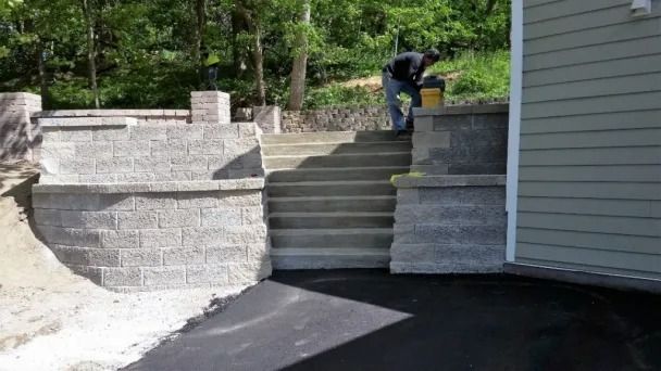 A person works near concrete stairs leading up to a stone retaining wall next to a house with beige siding.