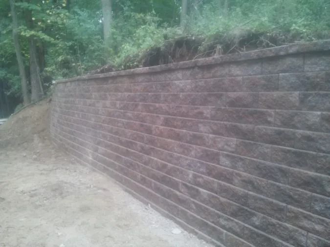 A reddish-brown stone retaining wall set against a dirt embankment and a forest background.