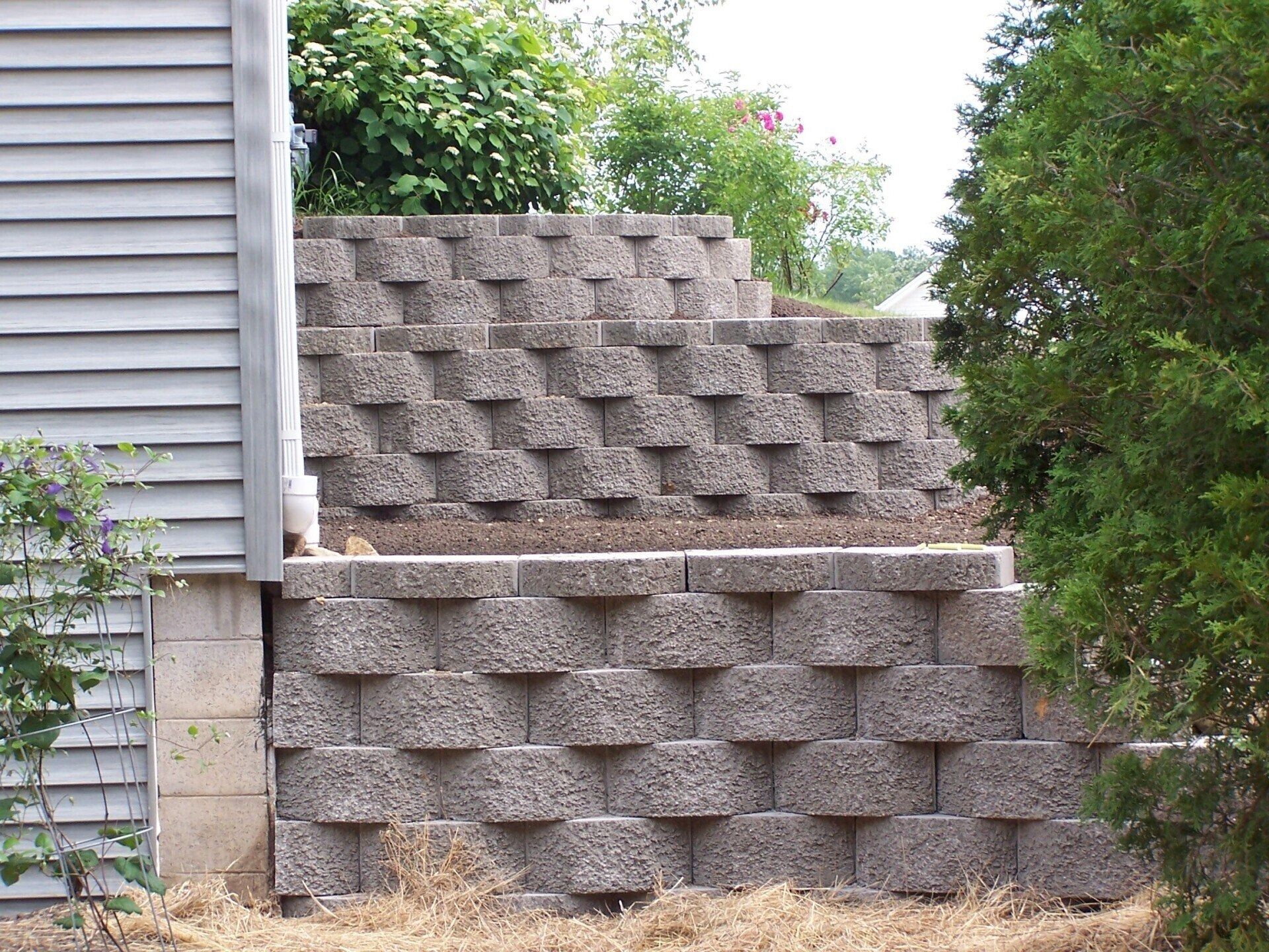 A multi-tiered retaining wall made of stacked grey concrete blocks sits alongside the siding of a residential house.