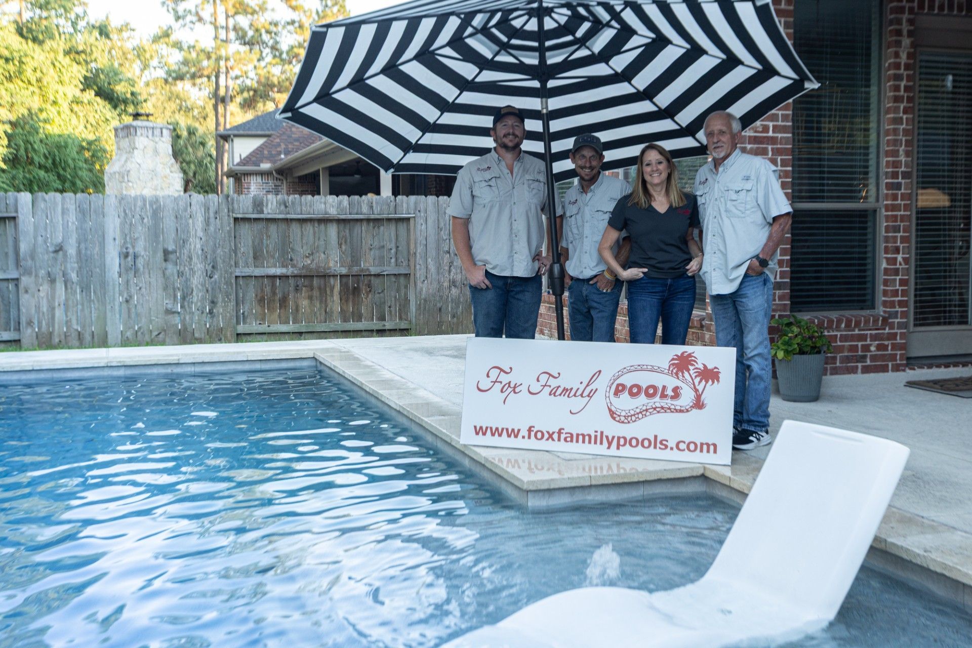 Four people stand by a pool, under a striped umbrella.