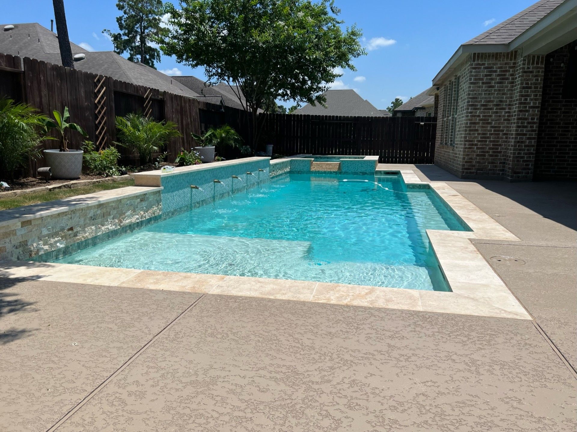 Pool with turquoise water, beige coping, and textured concrete patio, surrounded by a fence and brick house.