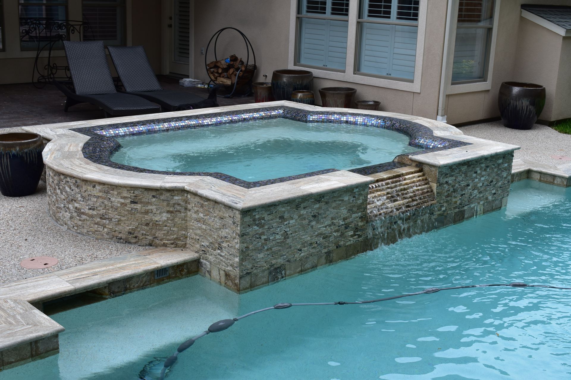 Outdoor swimming pool with attached hot tub, water feature, and patio with lounge chairs.