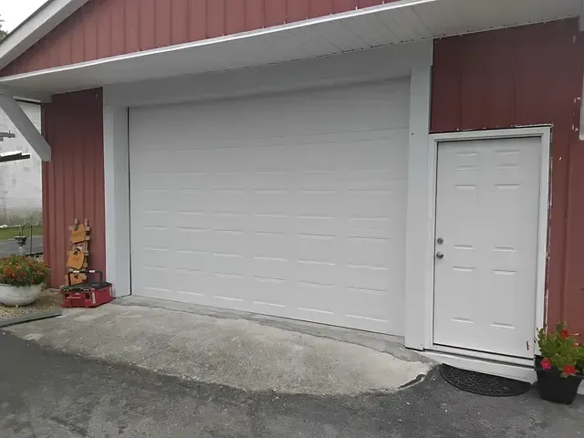 White garage door and matching entry door on a red building. Concrete ramp leads up to the garage.