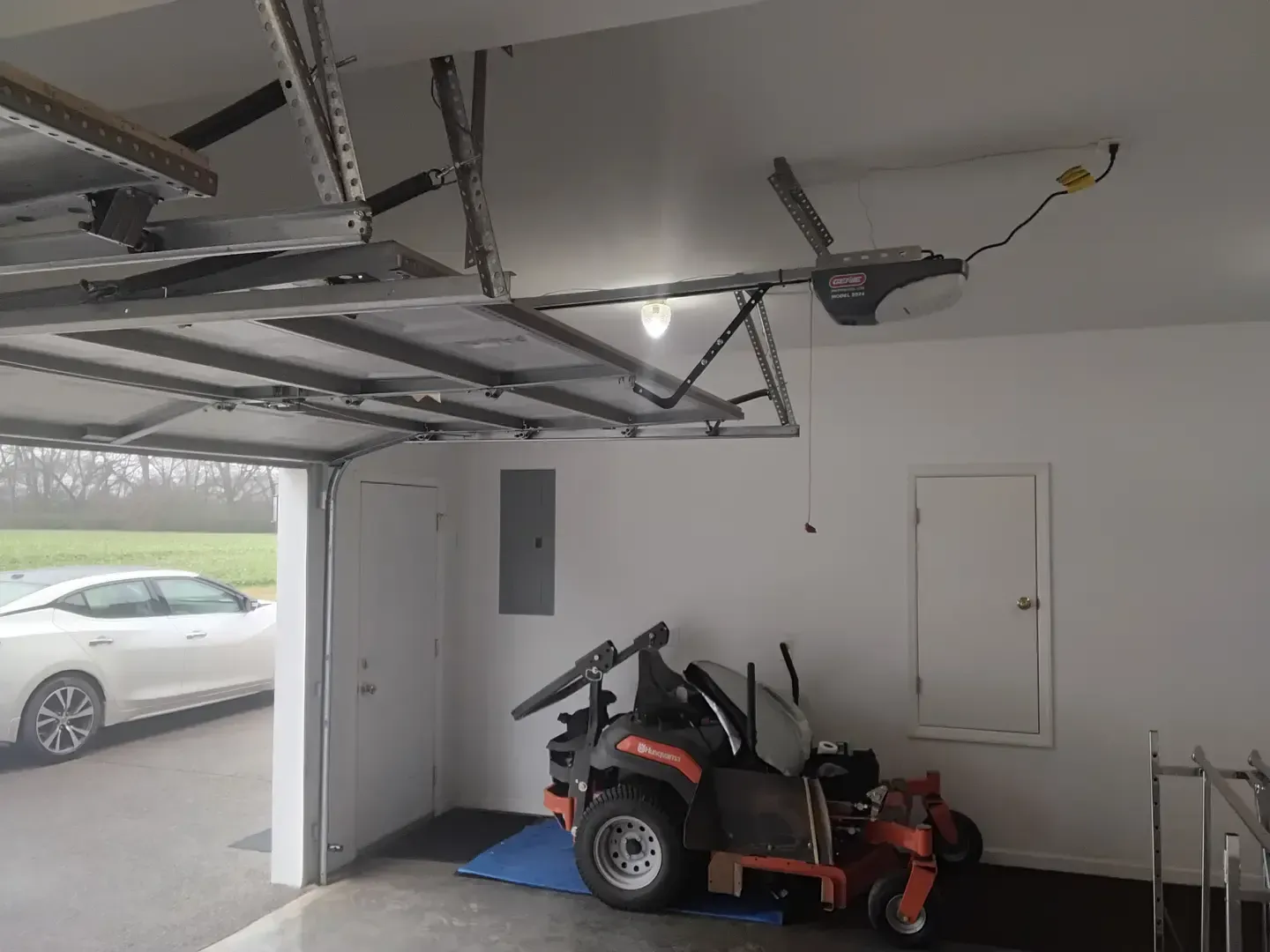 Garage interior with a partially open door, lawnmower, car visible, and garage door opener.