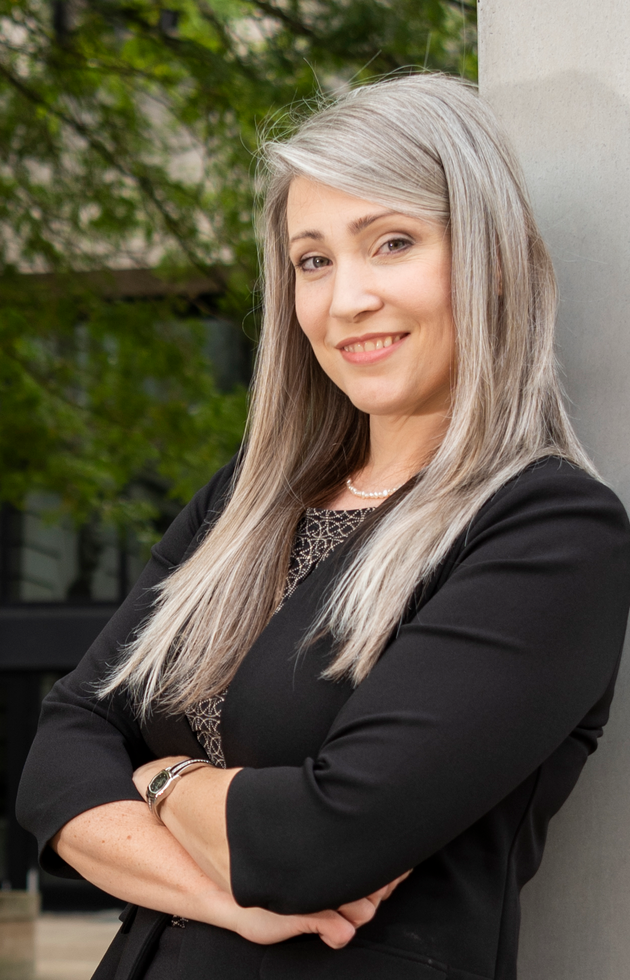 Smiling person with long gray hair and crossed arms, wearing a black blazer, standing outdoors.