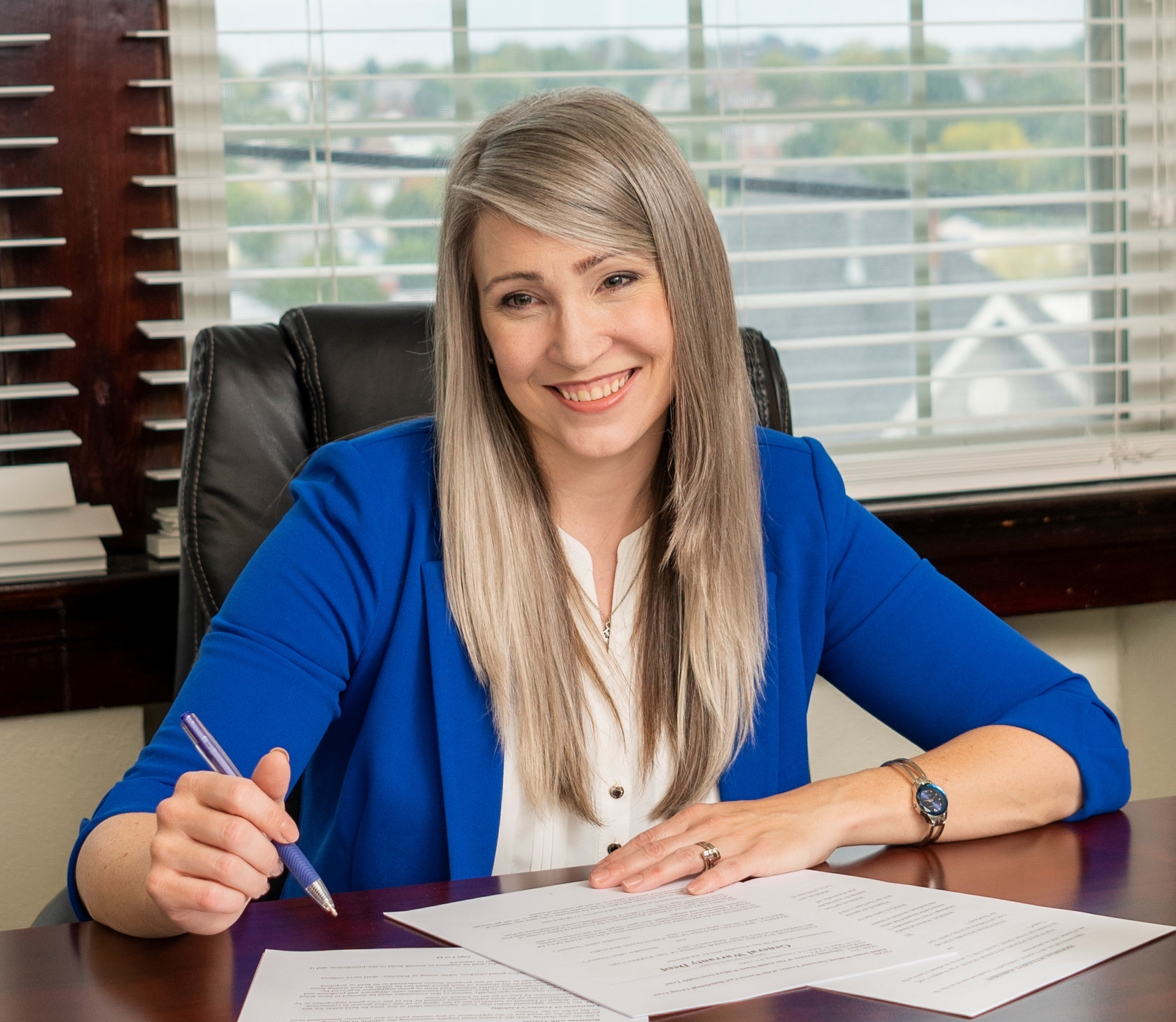 Smiling woman in a blue blazer writing at a desk with papers in an office.