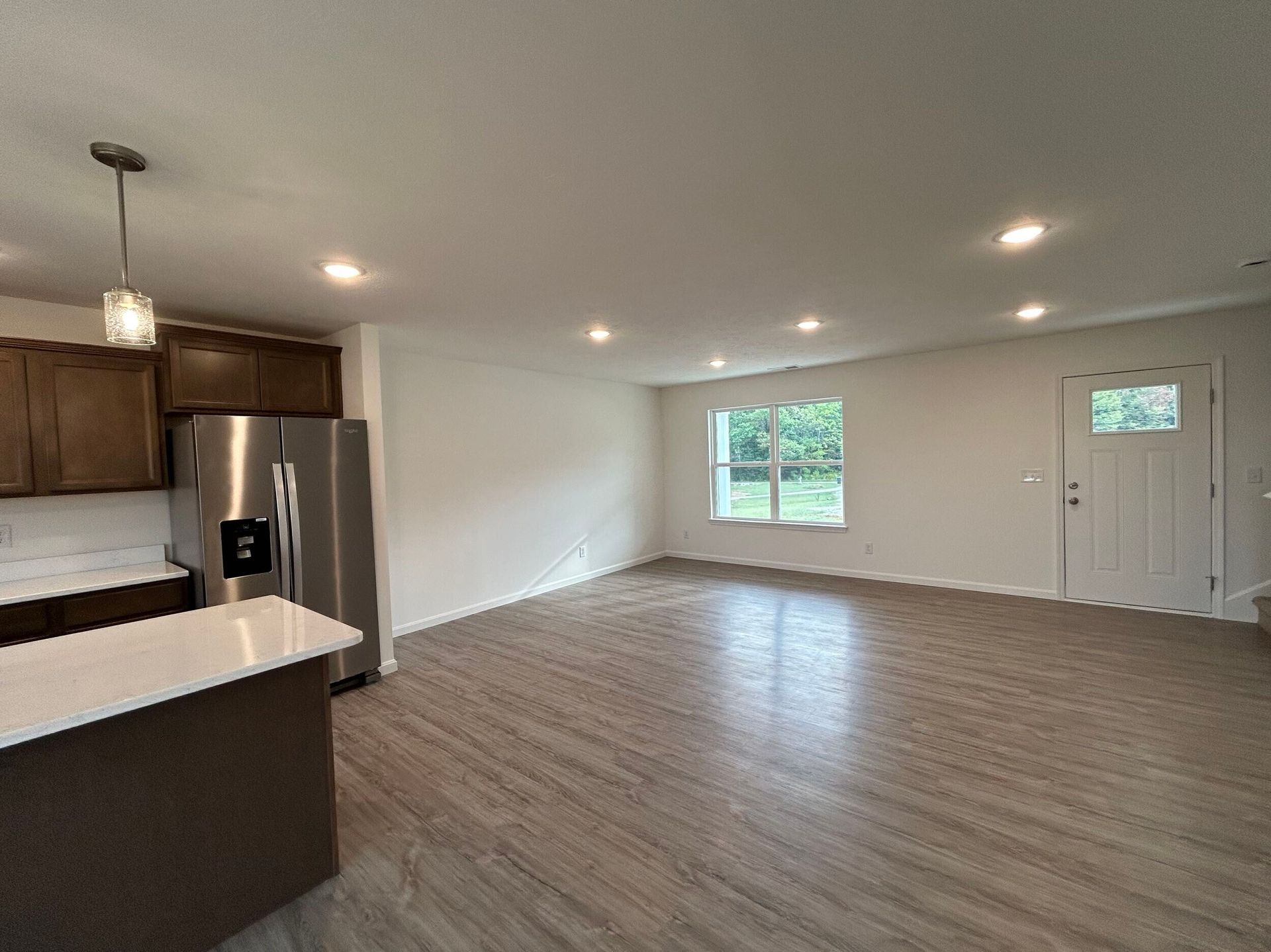 Empty open-plan kitchen and living area with white walls, wood floors, stainless refrigerator, and front door windows
