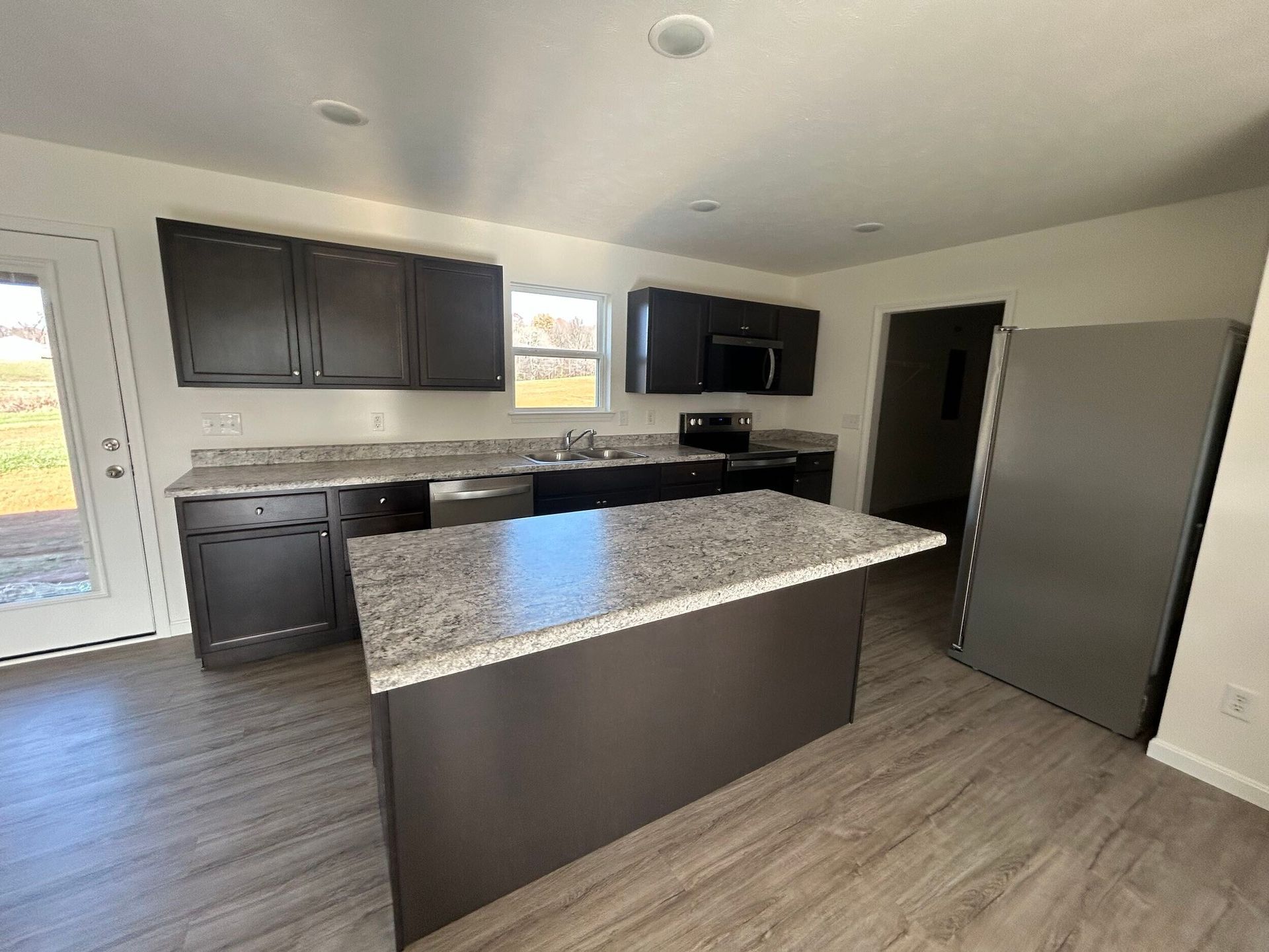 Modern kitchen with dark cabinets, gray island countertop, stainless fridge, and light wood floors