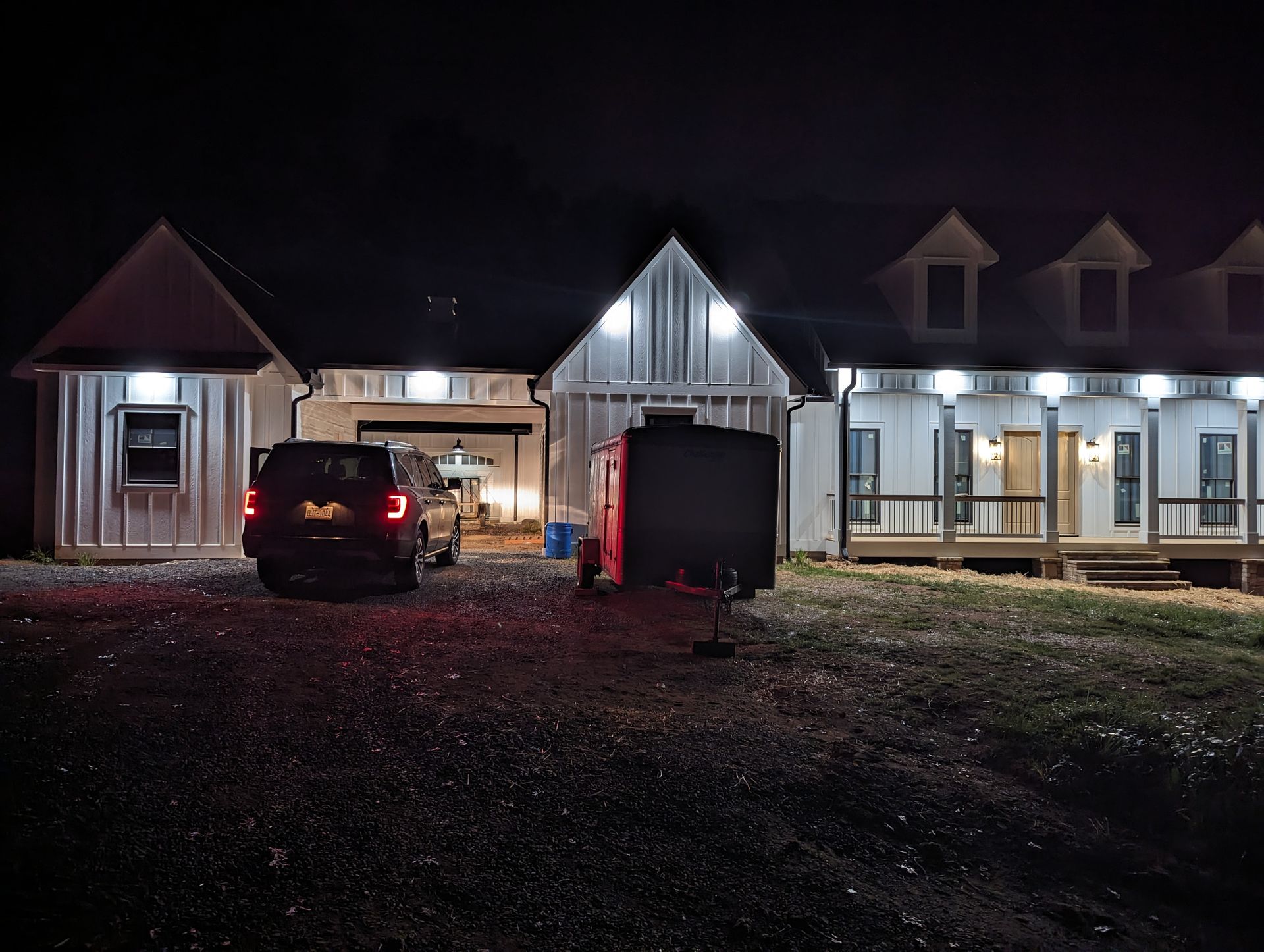 Night view of a house with attached garage and detached building. A car and trailer are parked in the gravel driveway.