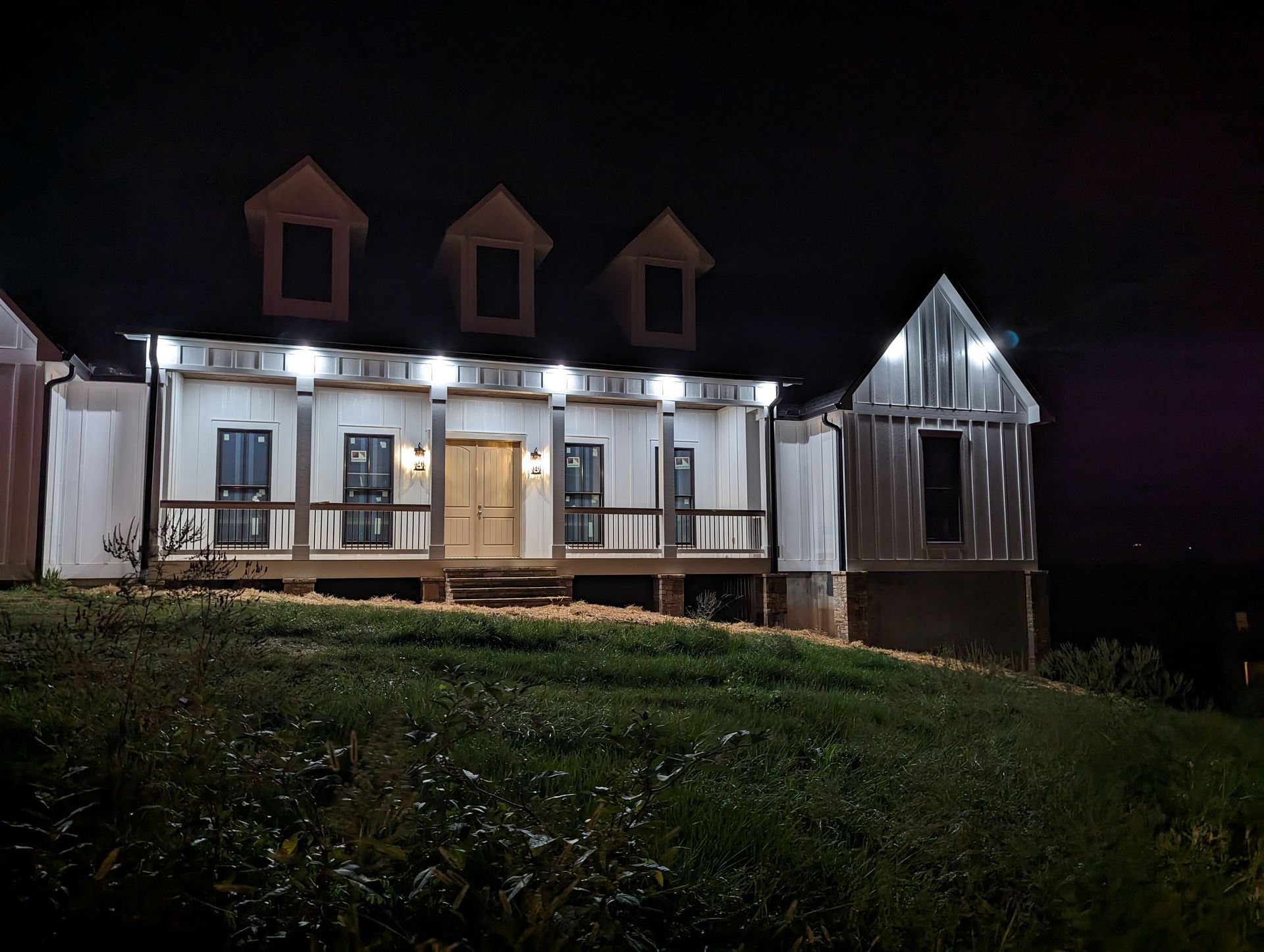Night view of a partially constructed white house with porch lights illuminating the front and a grassy hill.