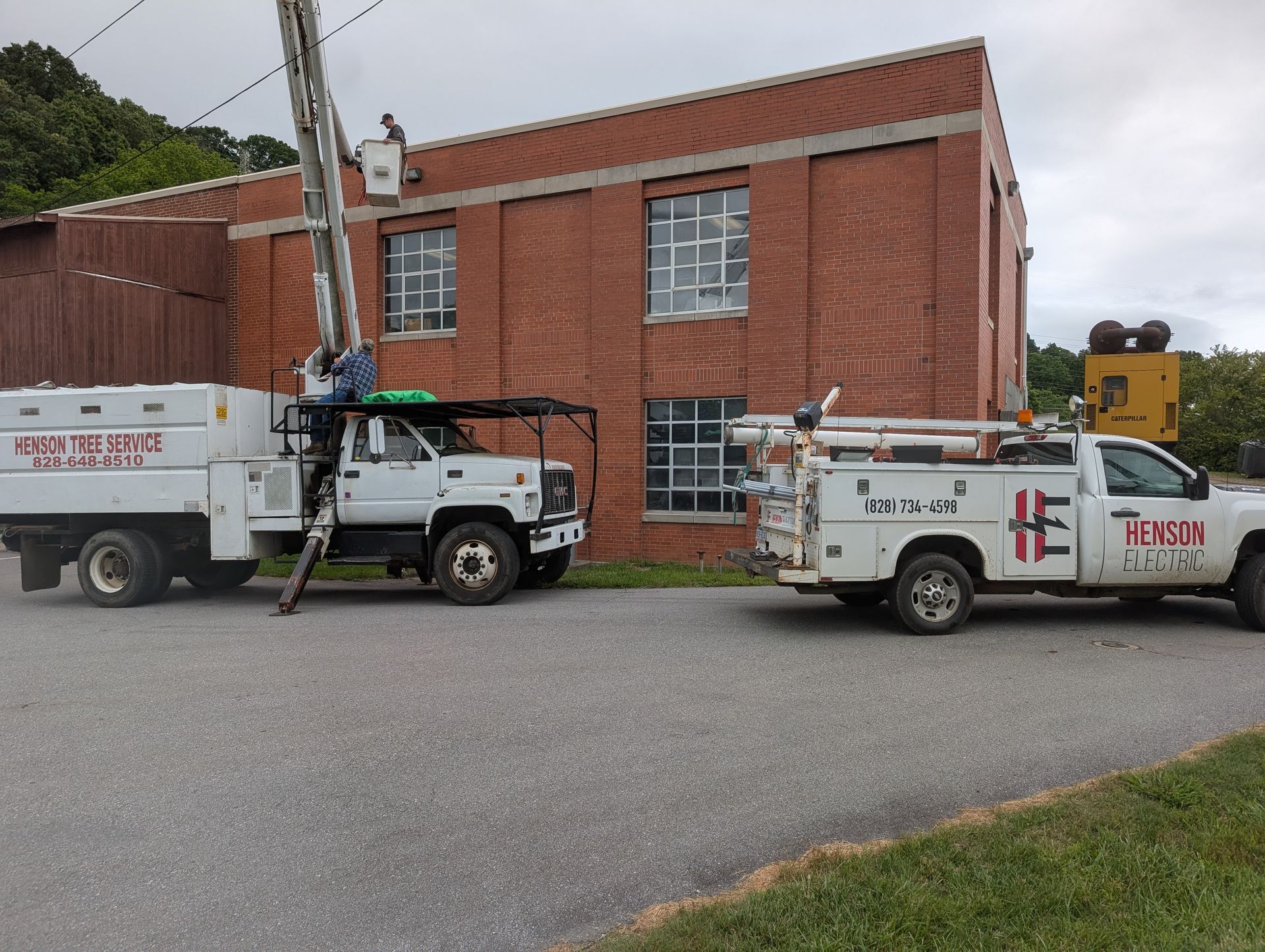 Two white utility trucks near a red brick building; one has a lifted boom.