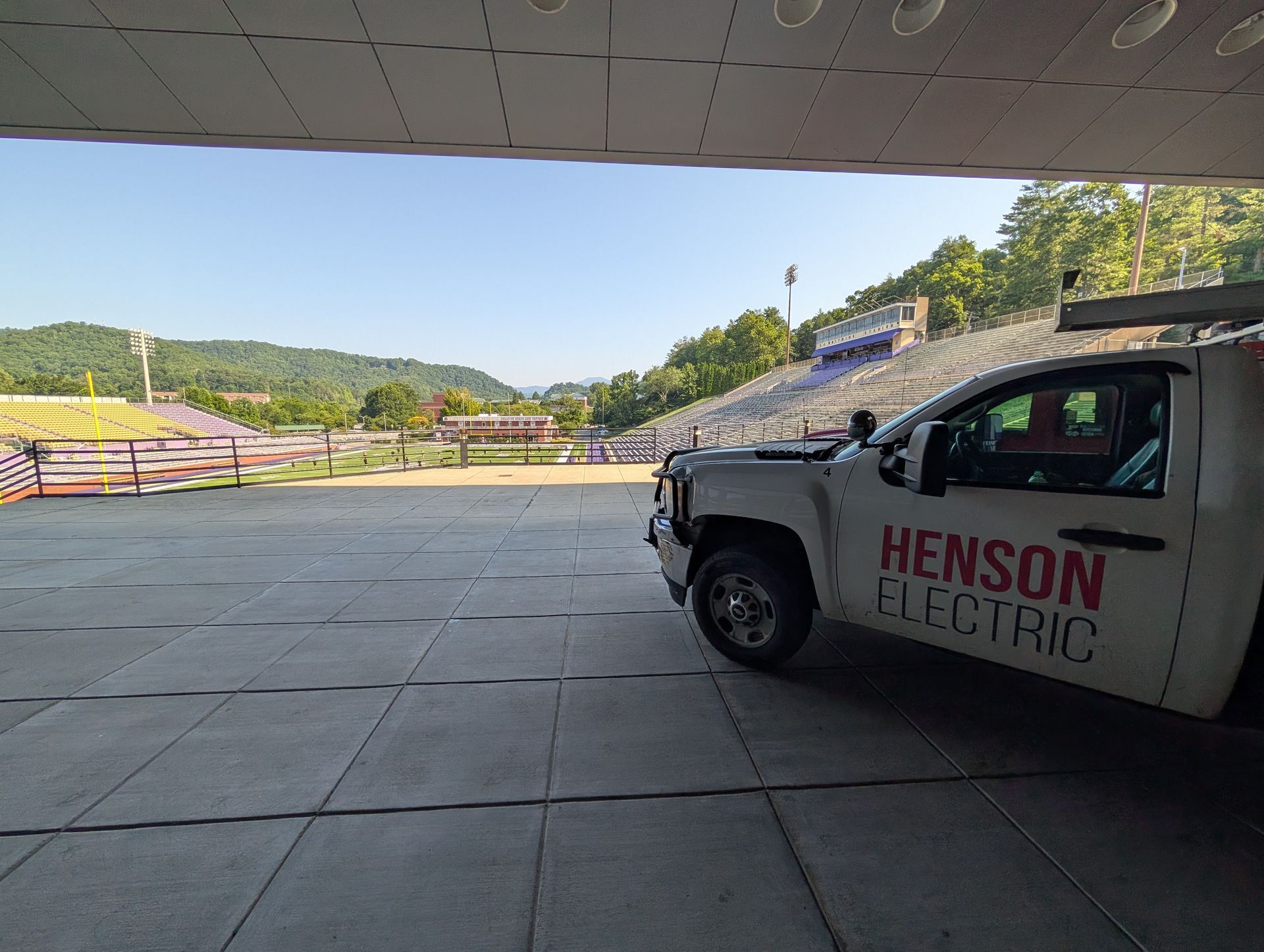 White Henson Electric truck parked under a covered area, stadium and mountains in the background under a blue sky.