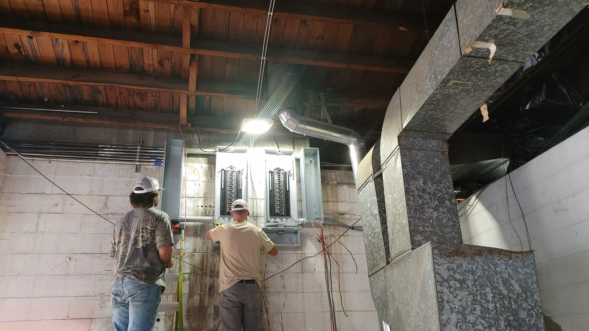 Two people working on electrical panel in a basement; ceiling and ducts visible.
