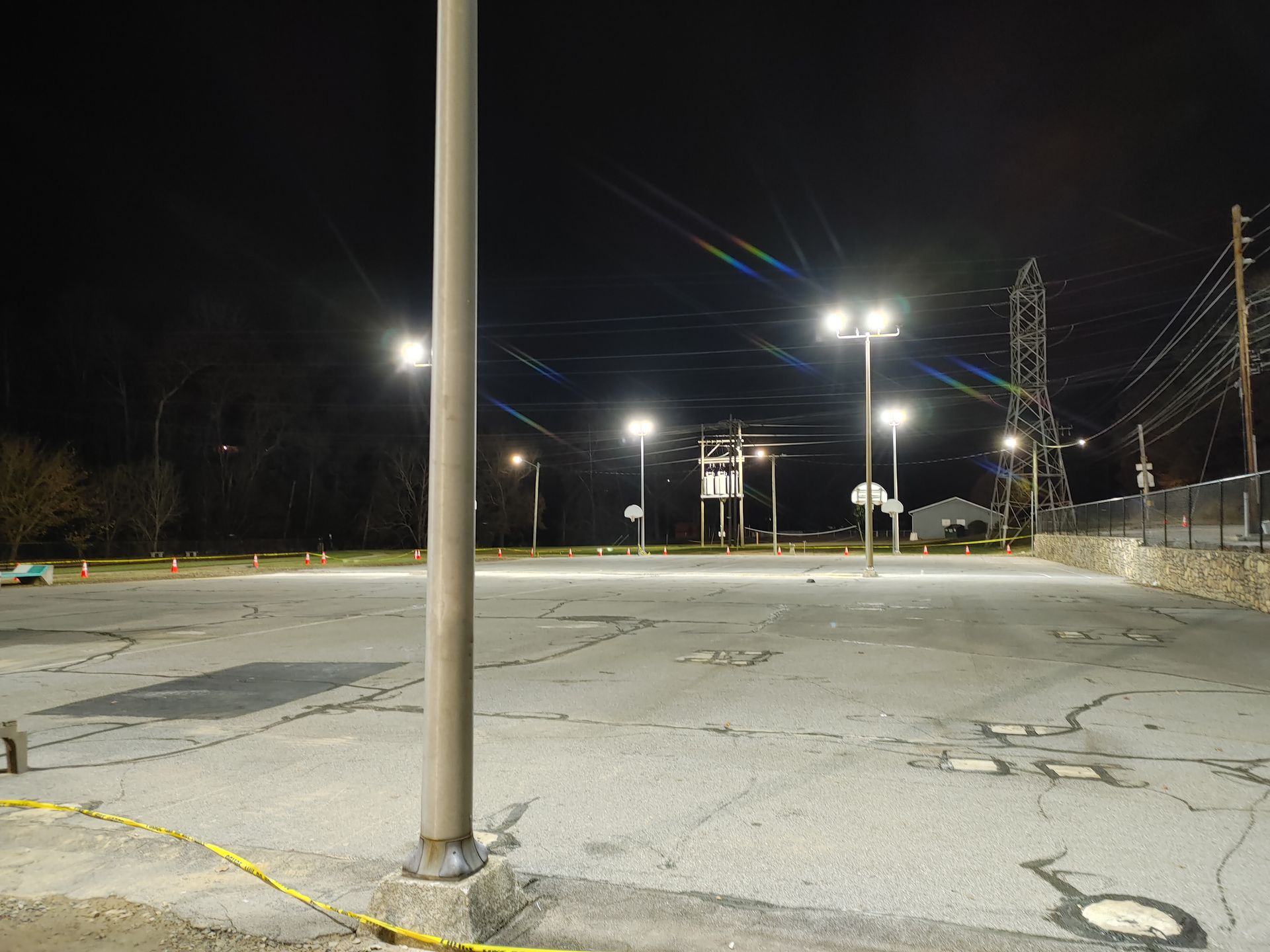 Nighttime view of an empty asphalt parking lot lit by tall streetlights. Basketball hoops are visible.