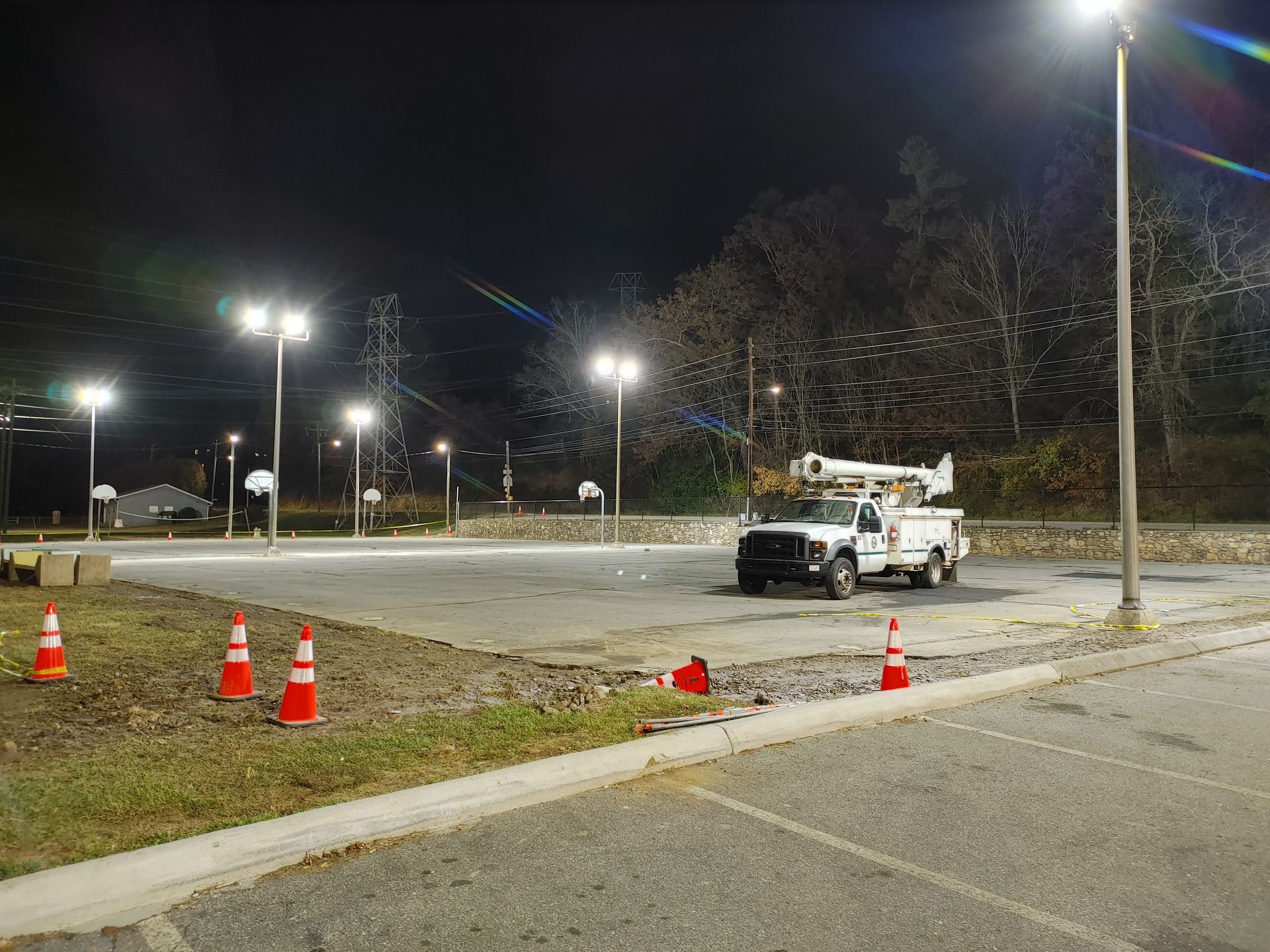 A utility truck at a basketball court lit by bright streetlights at night, surrounded by orange cones.