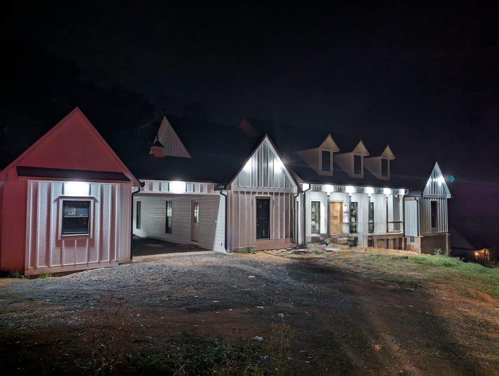House exterior at night, white siding, lighted windows, gravel driveway.