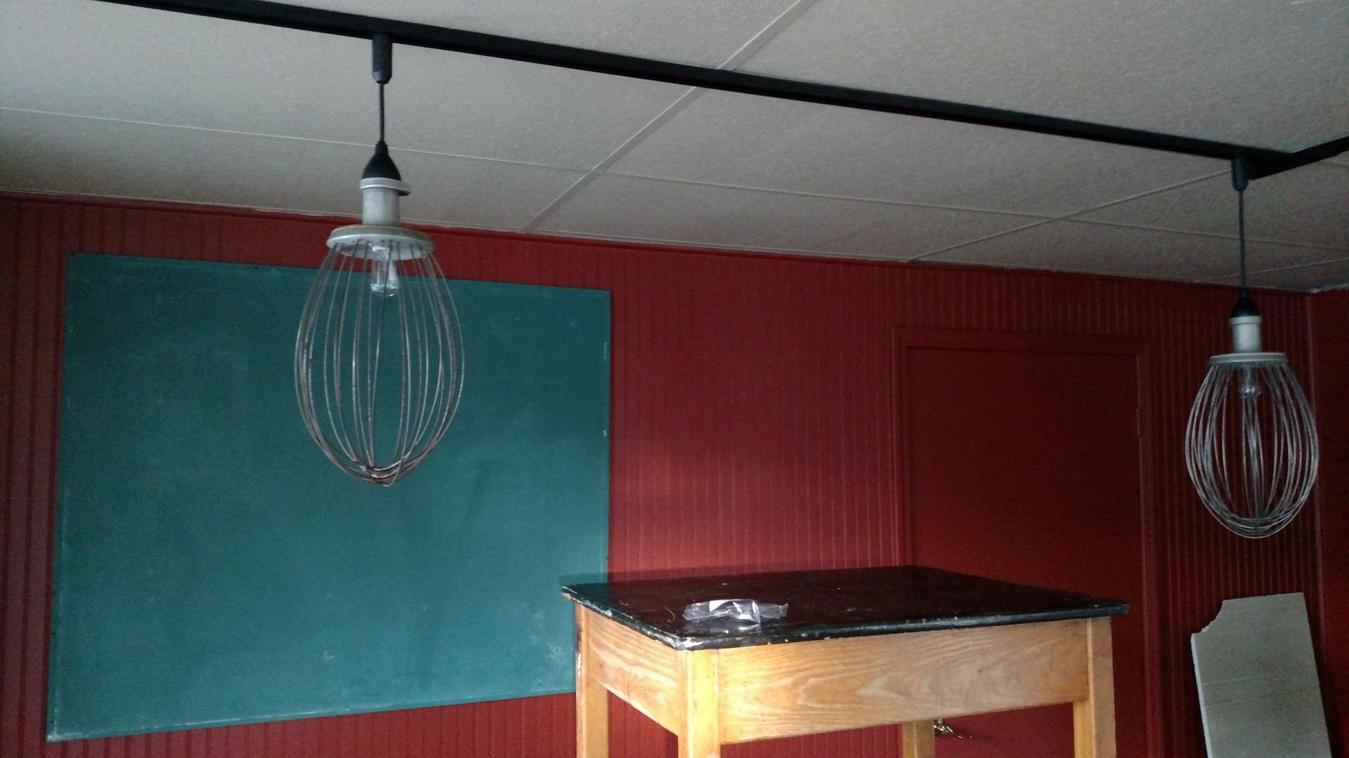 Two whisk-shaped pendant lights hang from a black track lighting system in a room with a chalkboard and red walls.