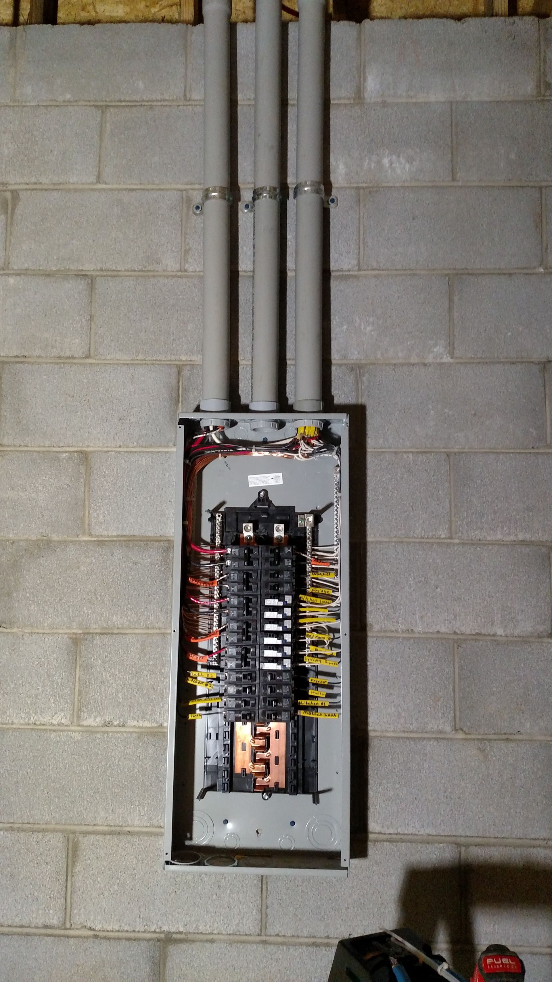 Electrical panel mounted on concrete block wall with conduits above.