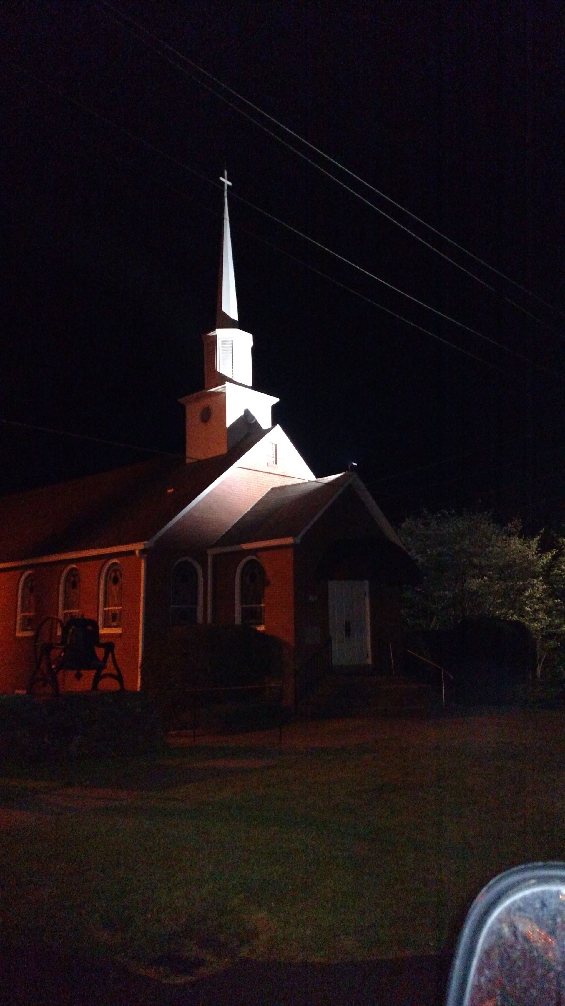 Church at night with steeple topped with a cross; illuminated facade against dark sky.