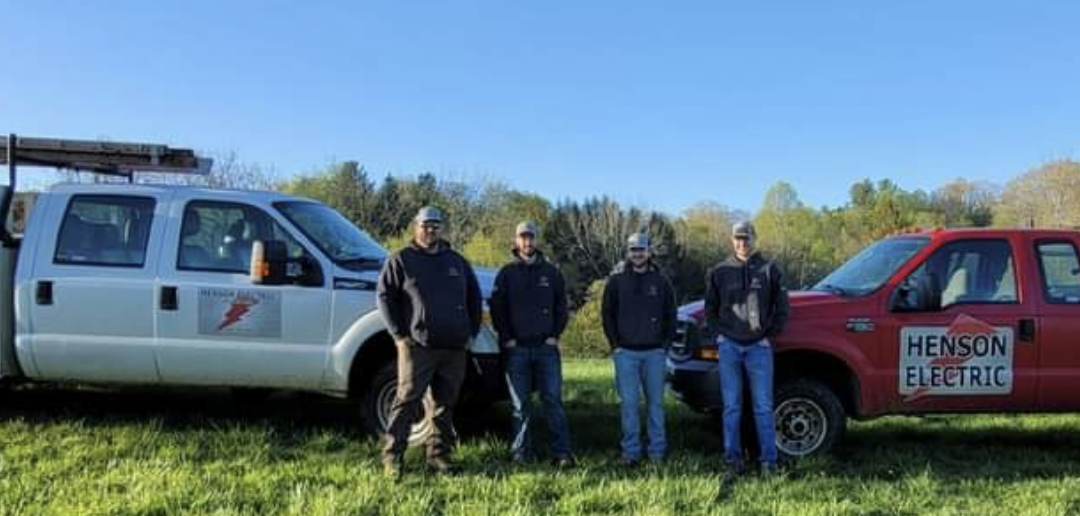 Four men in front of two work trucks on a grassy field. Clear sky. One truck is red, the other white.