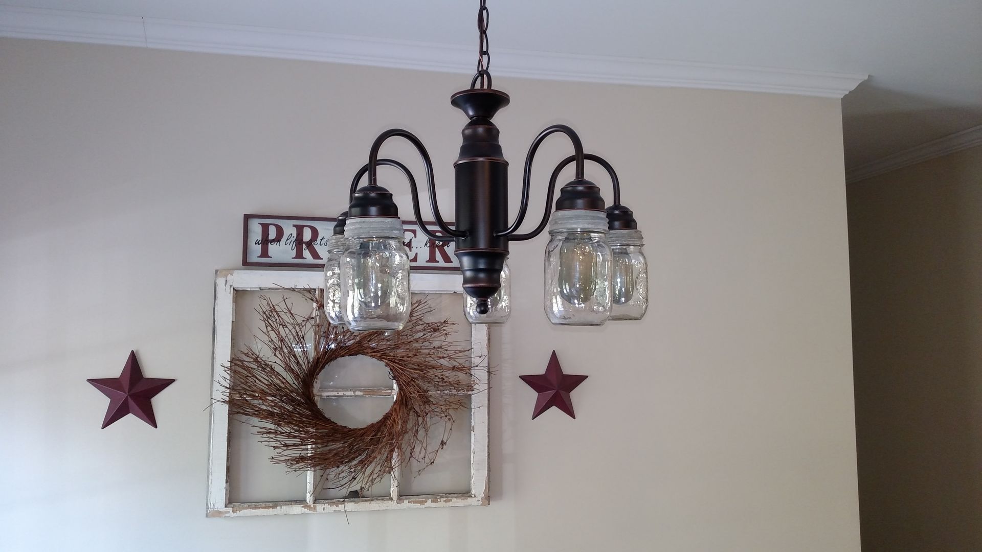Chandelier with mason jar shades hangs above farmhouse-style decor: window frame, wreath, and star accents.