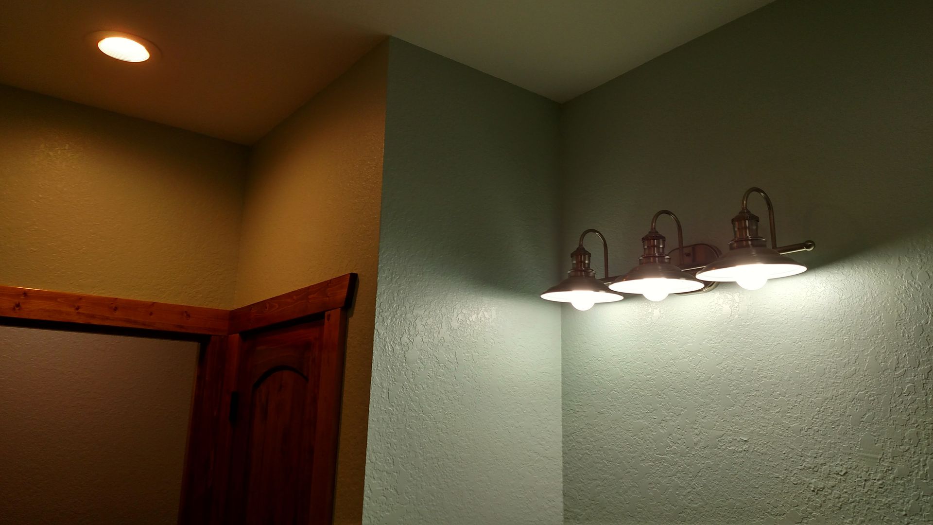 Bathroom corner with wall light fixtures and doorway. Light green walls, wooden door frame, overhead light.