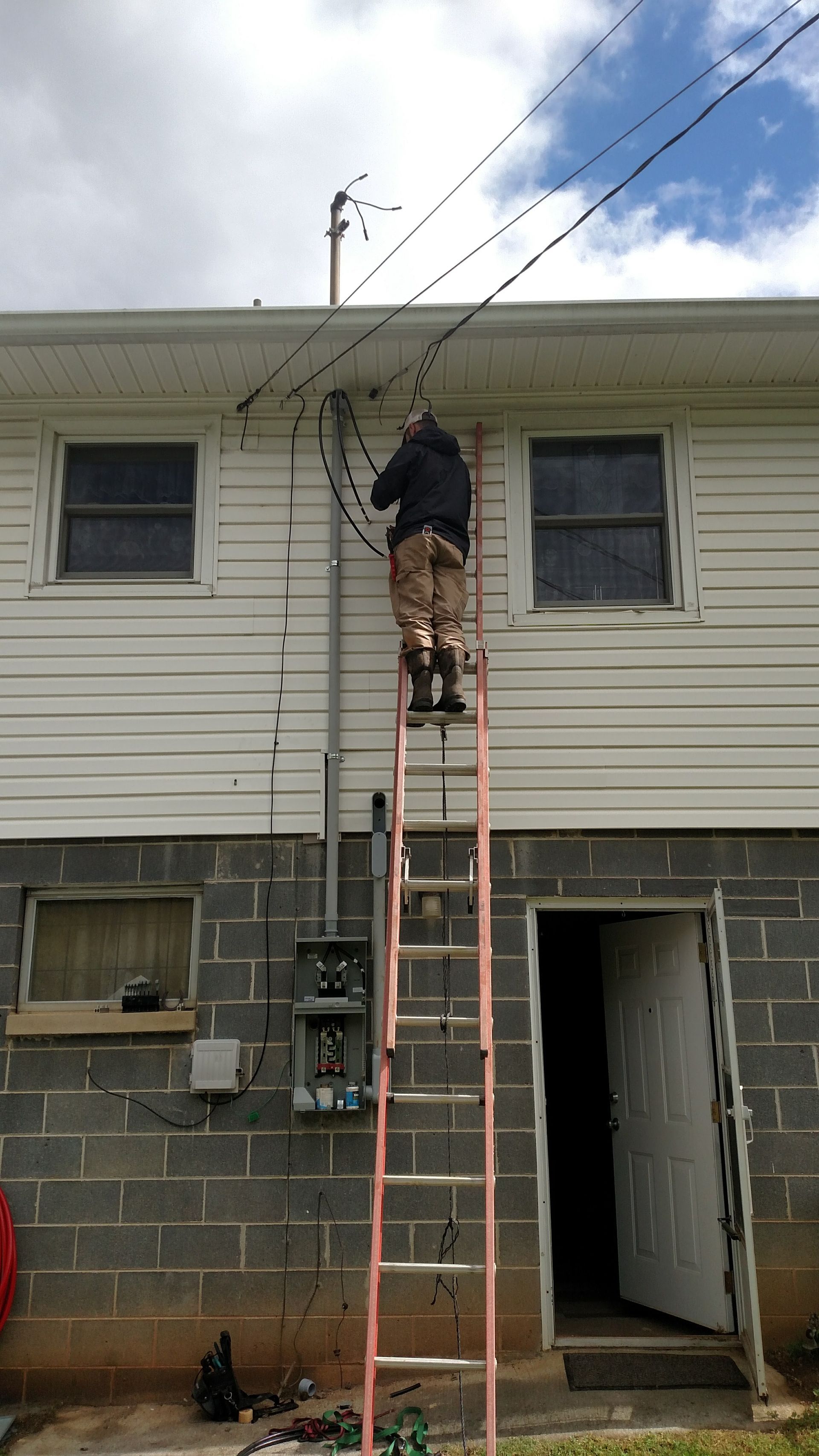 Person on a ladder working on overhead wires attached to a white house with dark windows. Gray brick foundation.