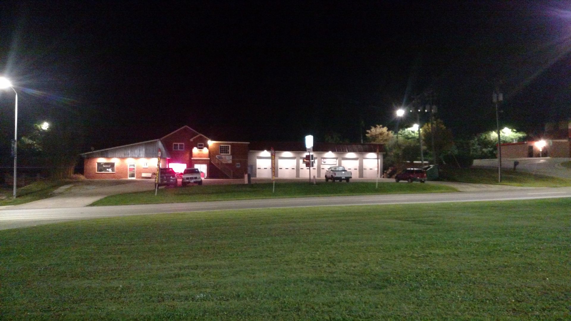 Nighttime view of a red-roofed building with several garage bays, cars parked in front.