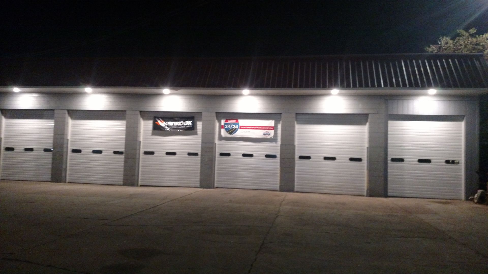 Exterior view of a commercial building with six garage doors, lit by overhead lights at night.