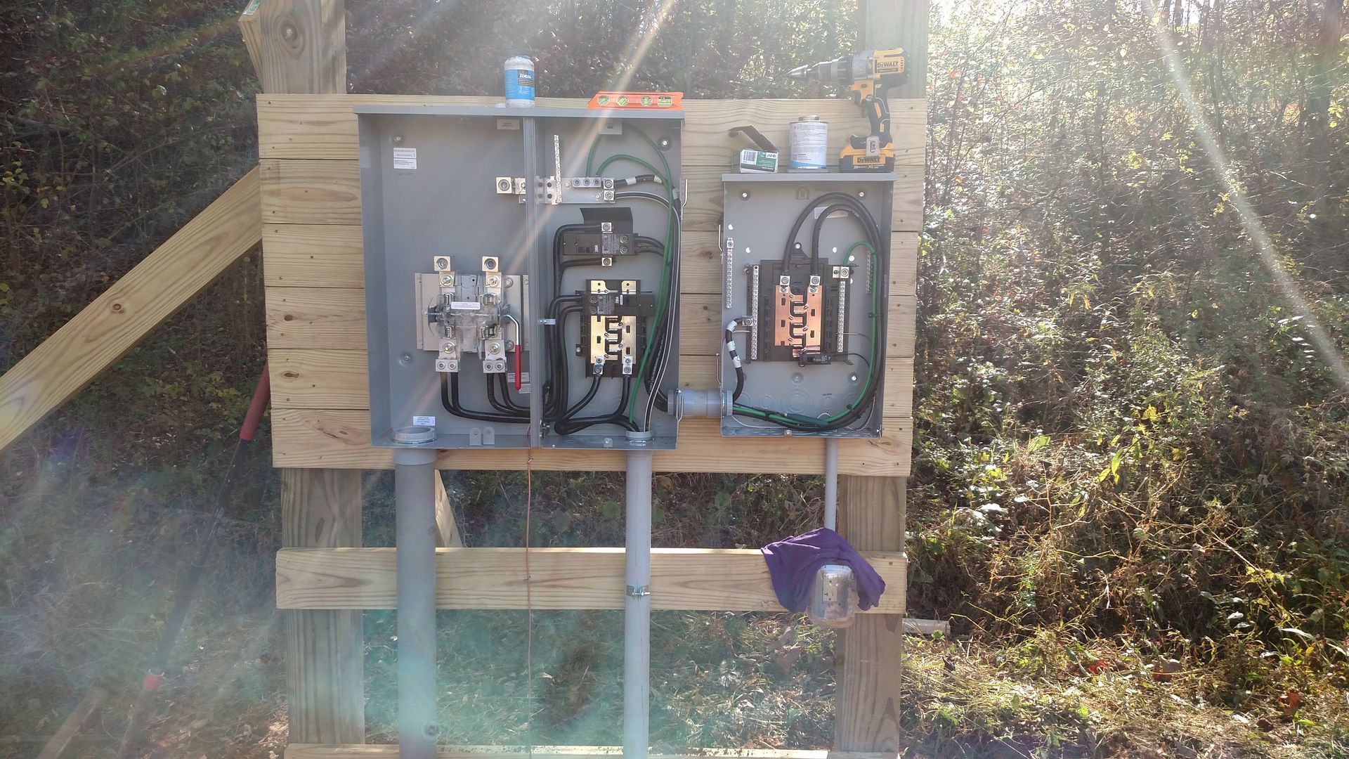 Electrical boxes mounted on a wooden board outdoors with conduits and wires visible. Bright sunlight.