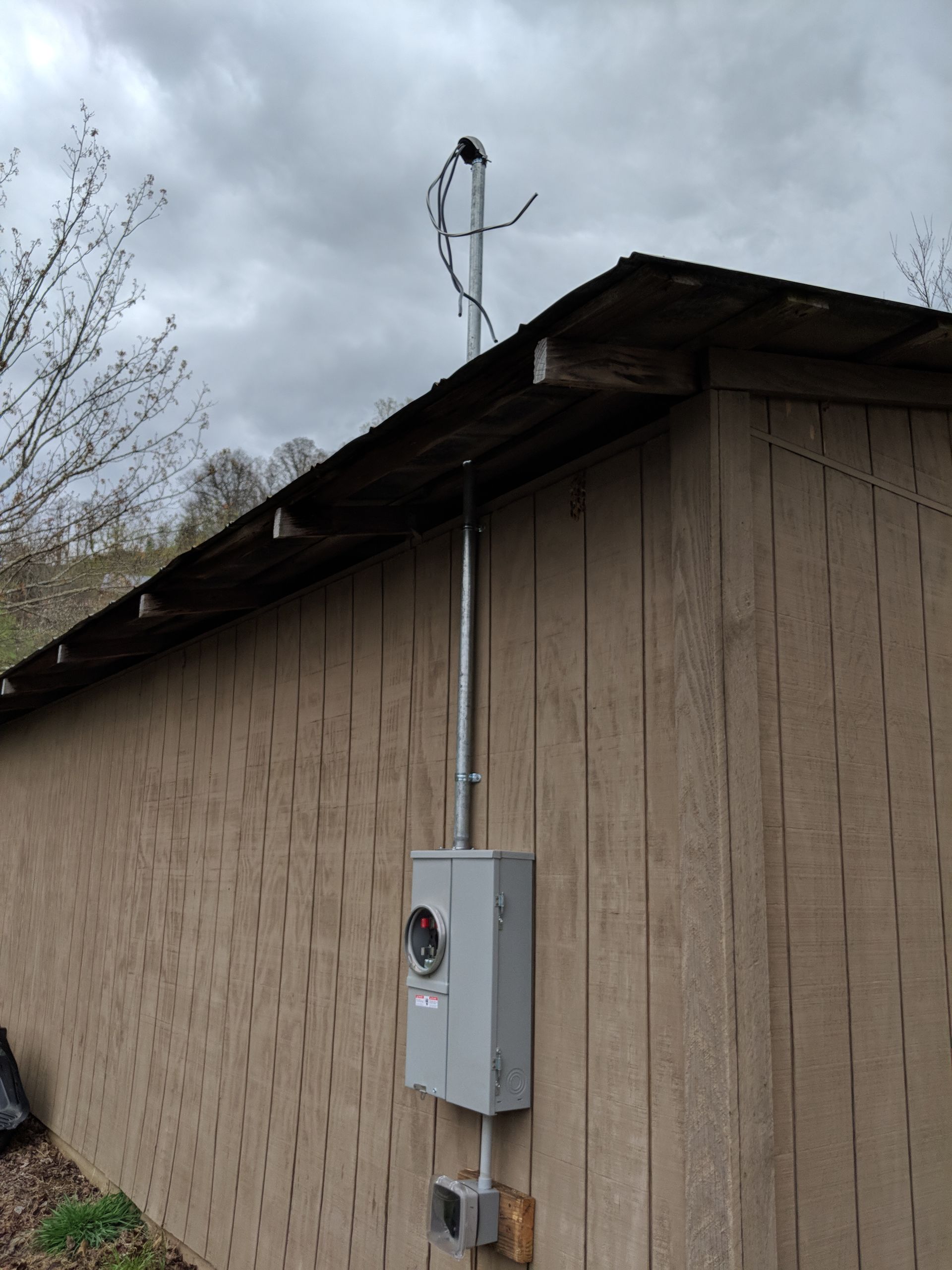 Electrical meter box on a wooden shed wall, with conduit running to roof and overhead wires; overcast sky.