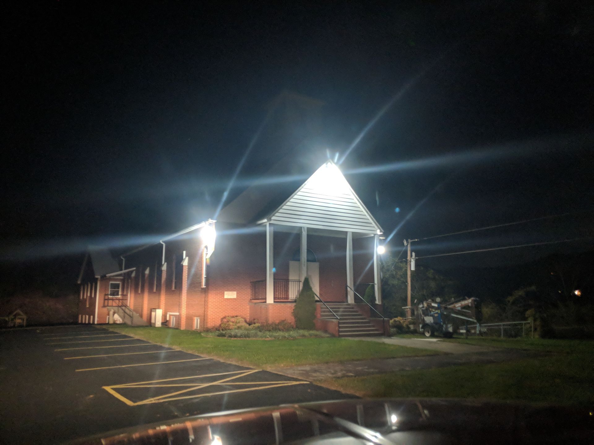 Brick building with a peaked roof illuminated at night, fronted by a parking lot and a porch with stairs.