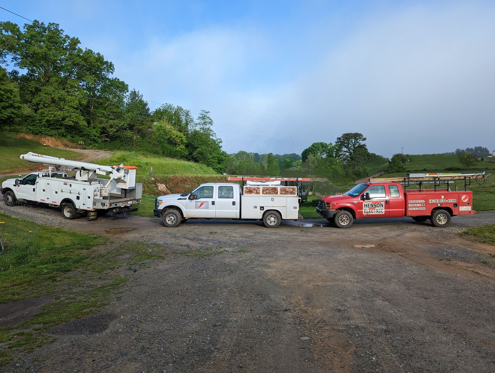Three work trucks parked on gravel, near trees. White, grey, and red vehicles.