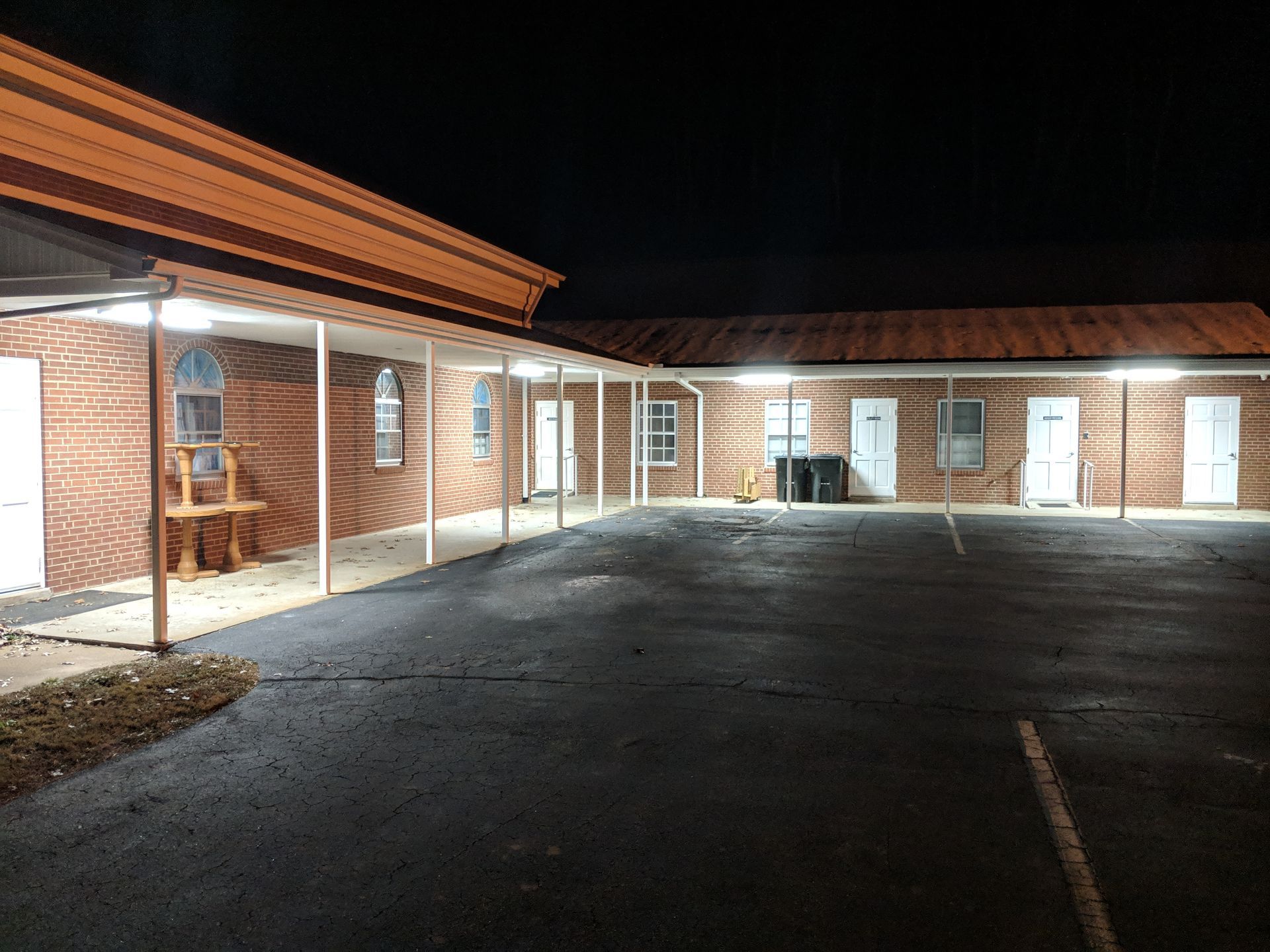 A brick building with an open walkway lit by overhead lights at night, with an empty parking lot.