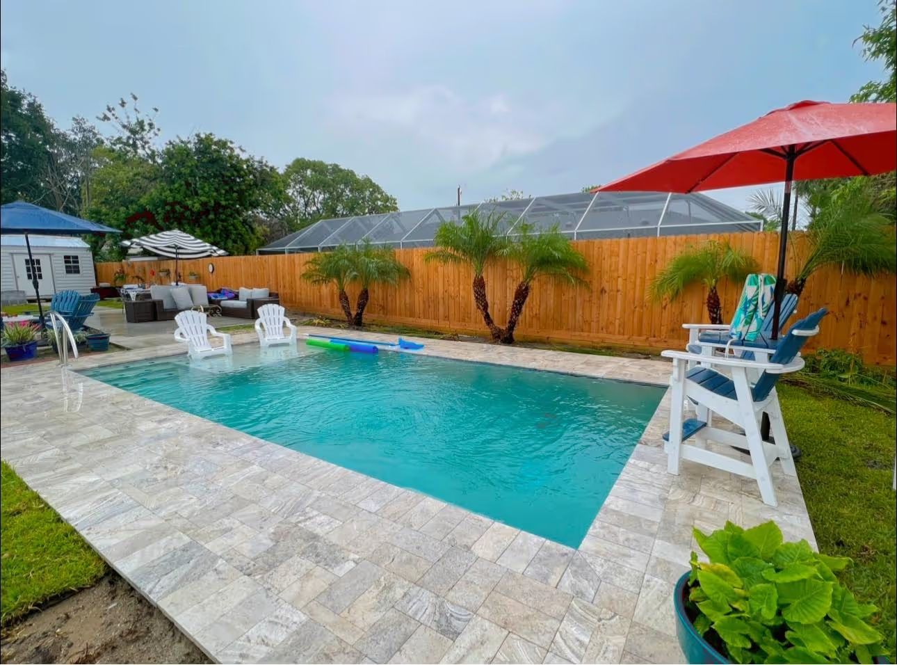 Backyard pool with turquoise water, white lounge chairs, red umbrella, and wooden fence under a cloudy sky