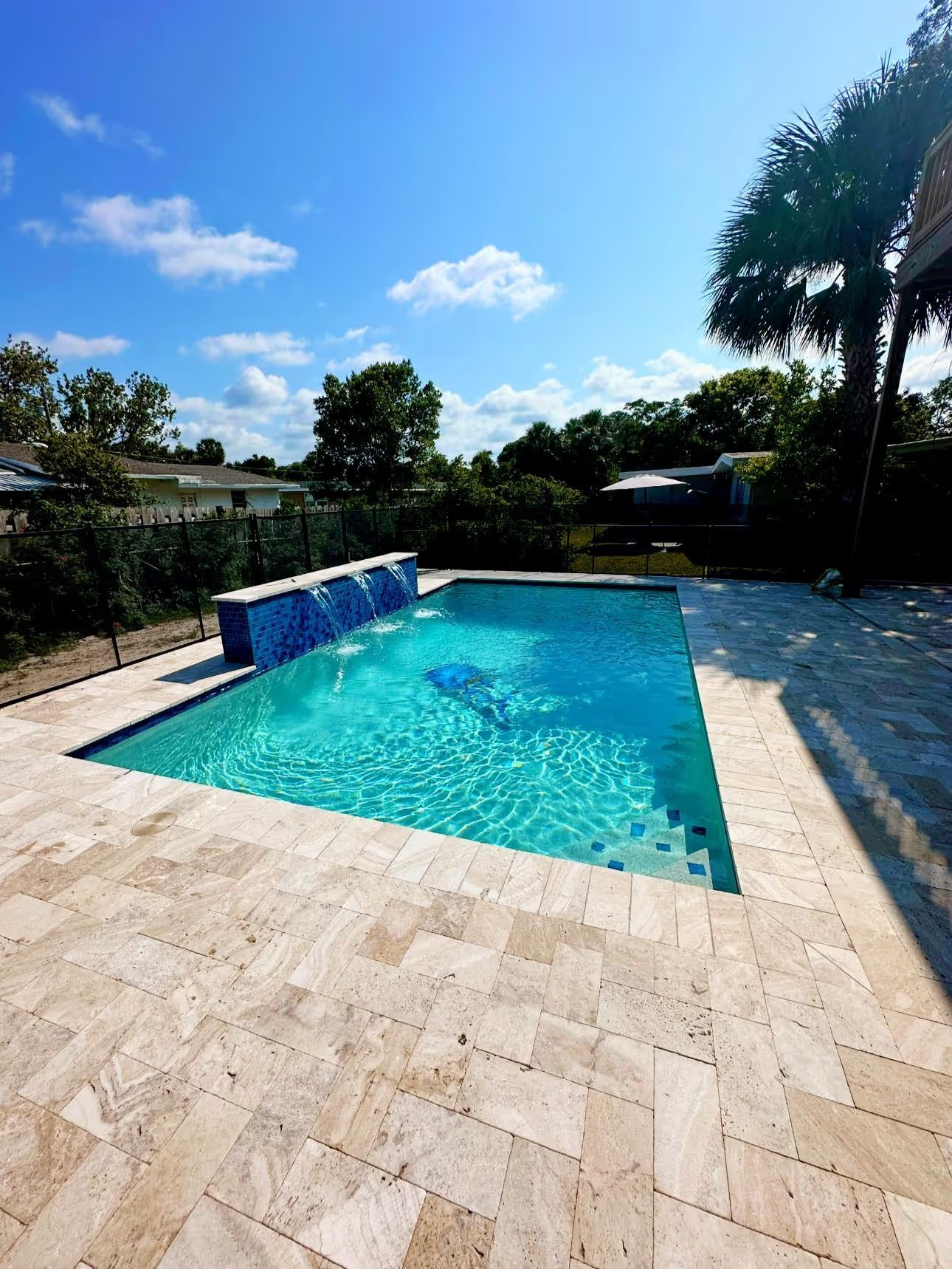 Bright turquoise backyard pool with stone deck, palm trees, and blue sky on a sunny day