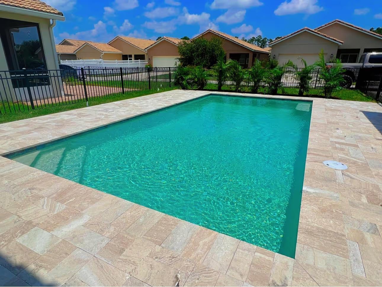 Rectangular turquoise swimming pool surrounded by beige stone patio in a suburban backyard