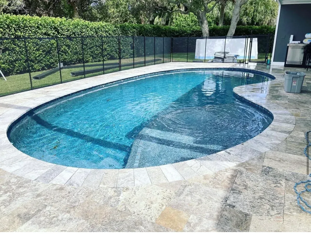Backyard swimming pool with blue water, stone deck, and black safety fence near greenery