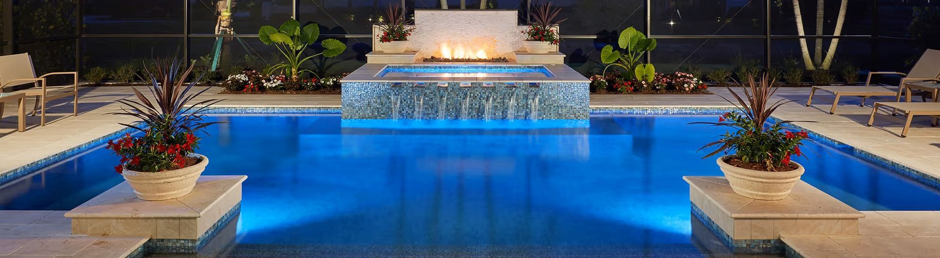 Illuminated outdoor pool with blue water, steps, and potted plants beside a lit fountain at night