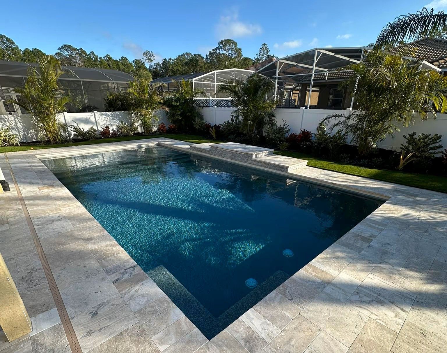 Rectangular blue backyard swimming pool surrounded by stone decking and greenery under a bright sky