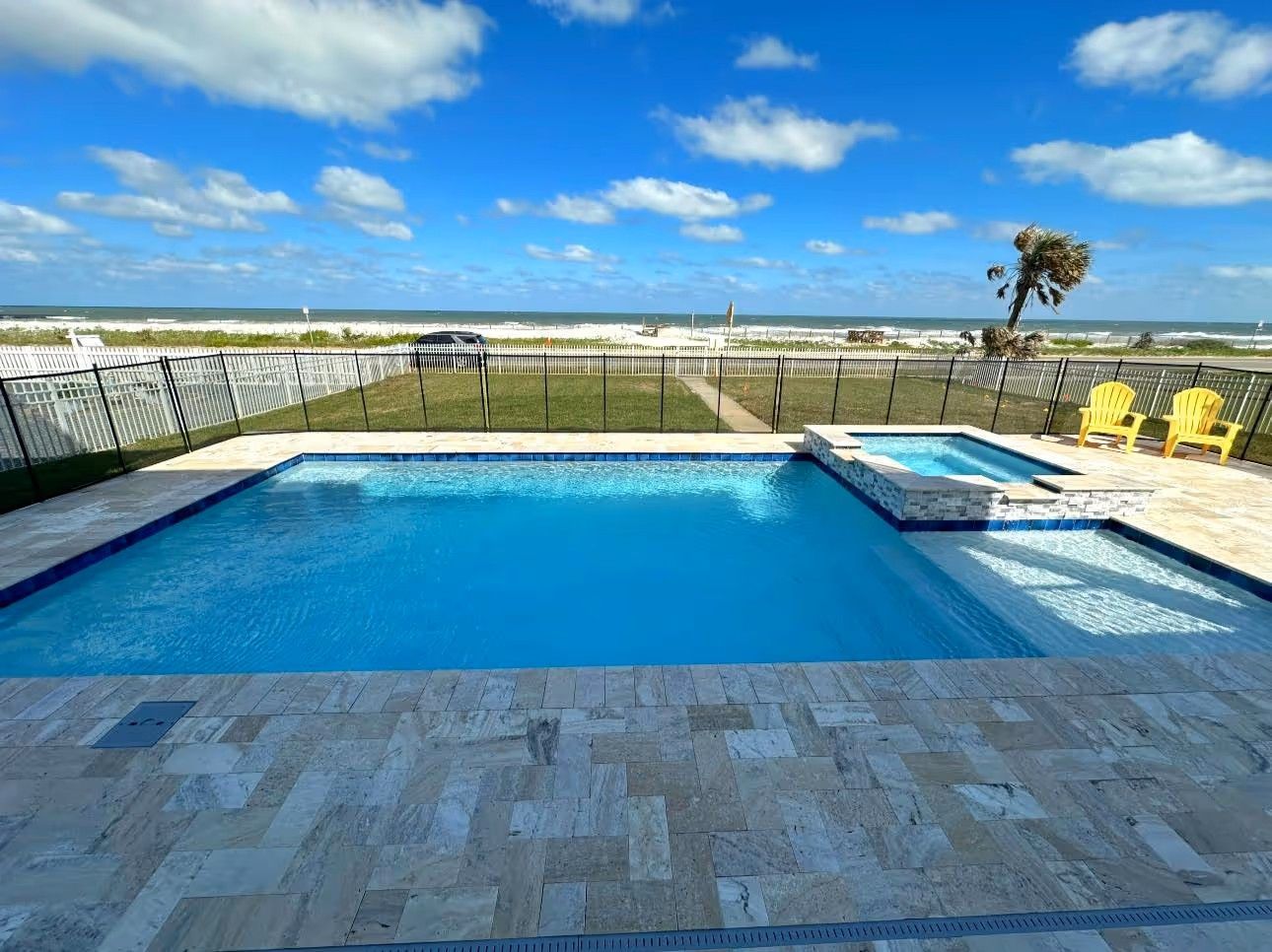 Oceanfront pool with blue water, tiled deck, and yellow lounge chairs under a sunny sky