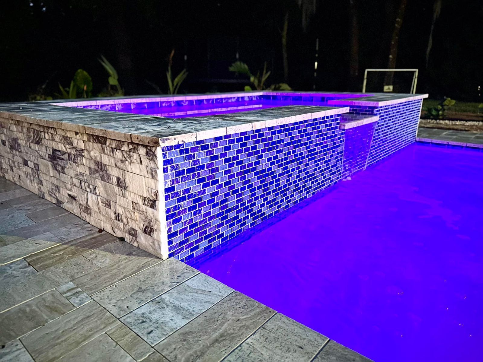 Nighttime pool with blue-purple lighting beside a stone-tiled deck and raised stone wall.