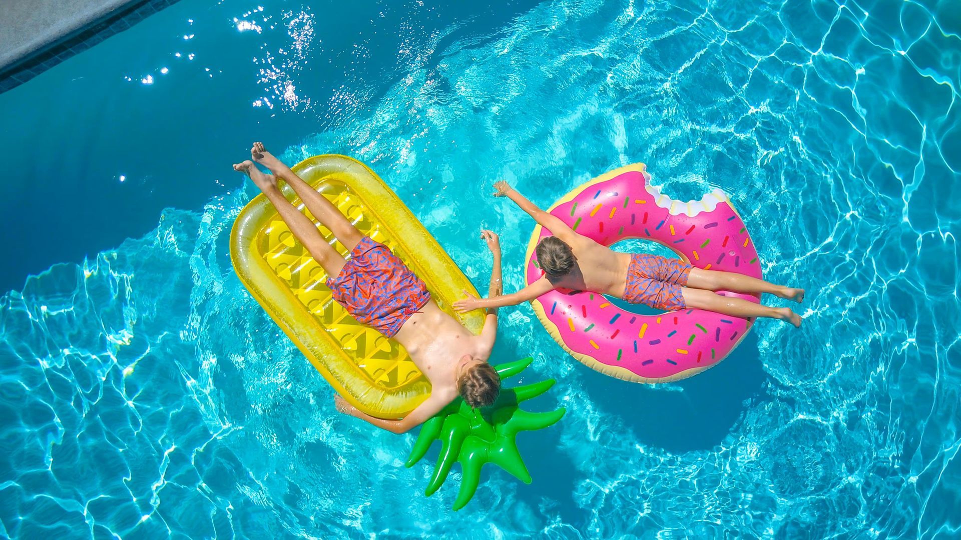Two people floating on pink and yellow inflatable rings in bright blue pool water