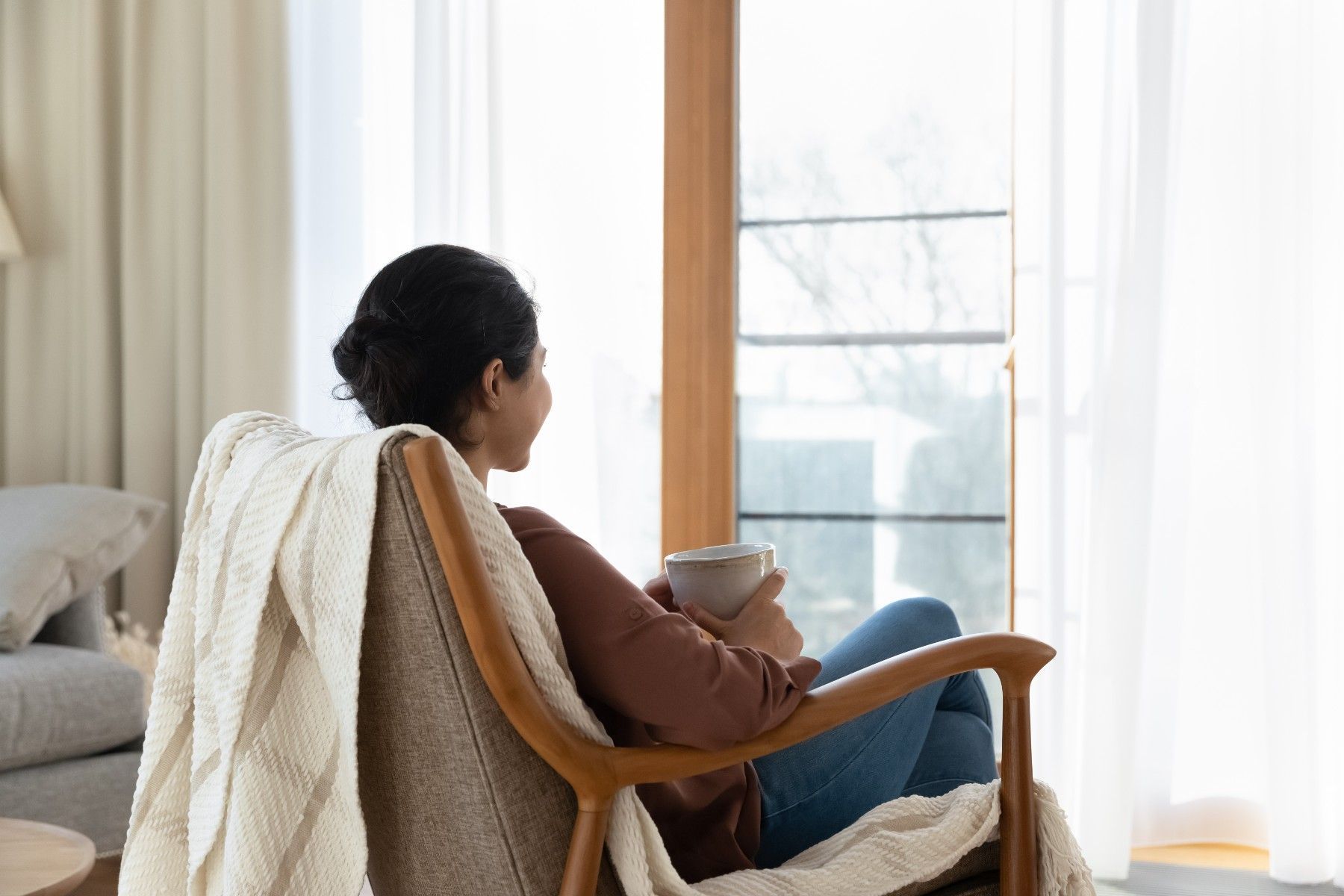 A woman is sitting in a chair holding a cup of coffee and looking out the window.