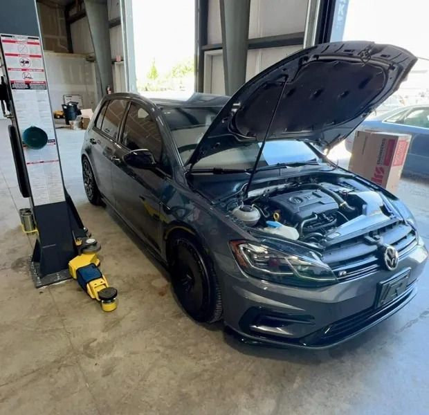 Gray Volkswagen Golf hatchback with hood open in a garage, next to a car lift.