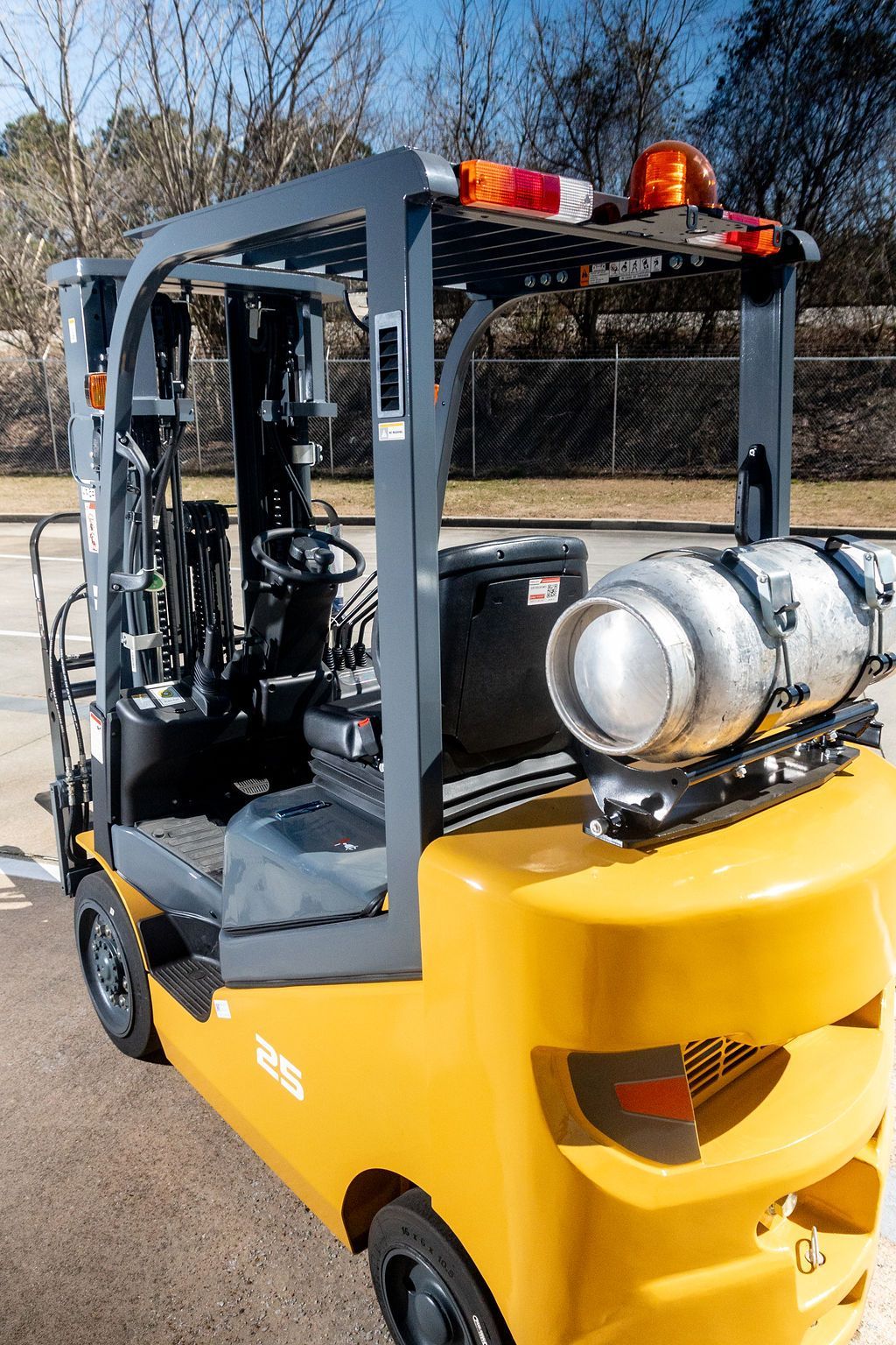 Yellow forklift with a propane tank and flashing lights parked outdoors near trees and a chain-link fence