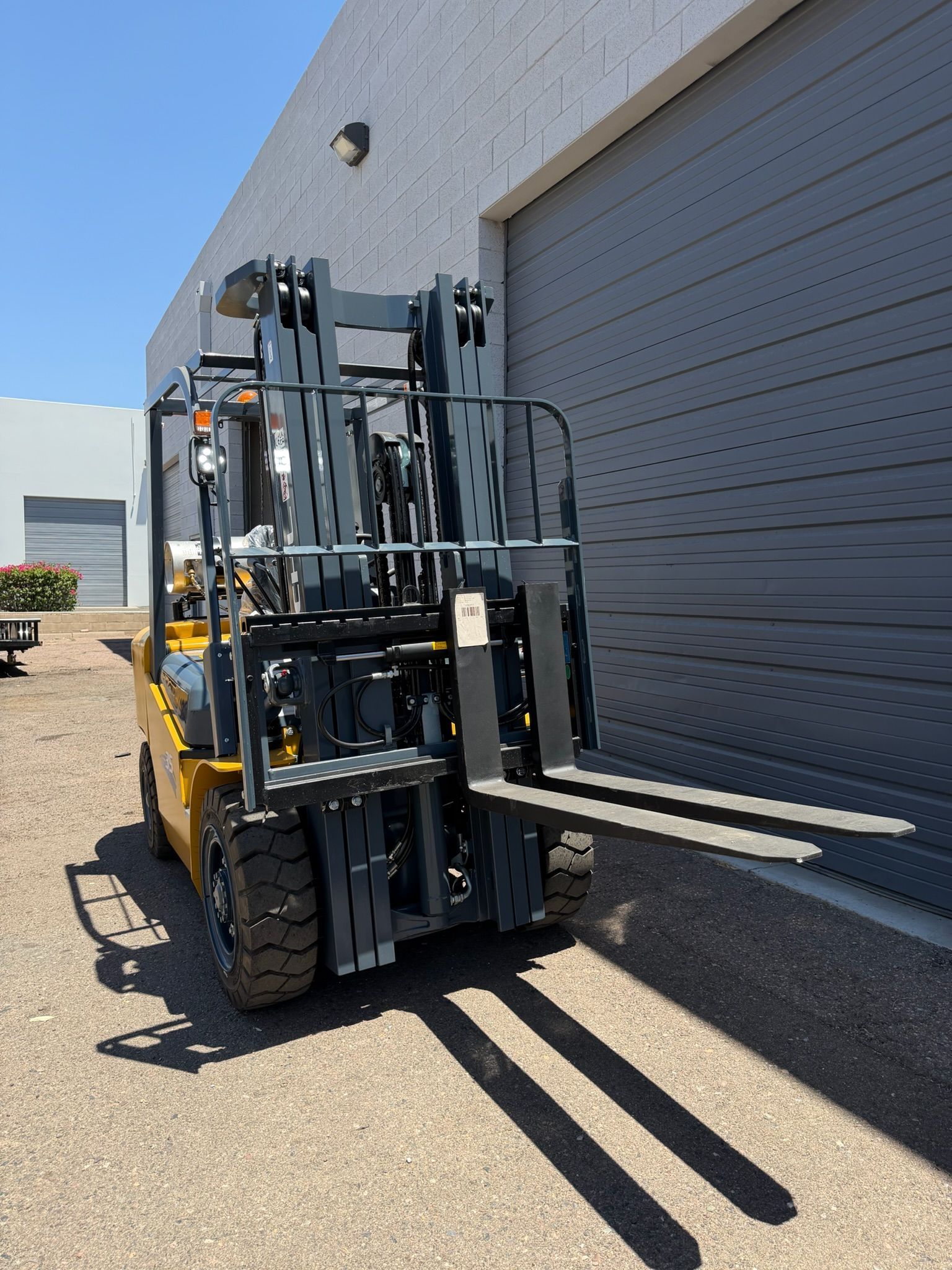 Yellow forklift parked beside a gray warehouse door, with forks raised in the sunlight.
