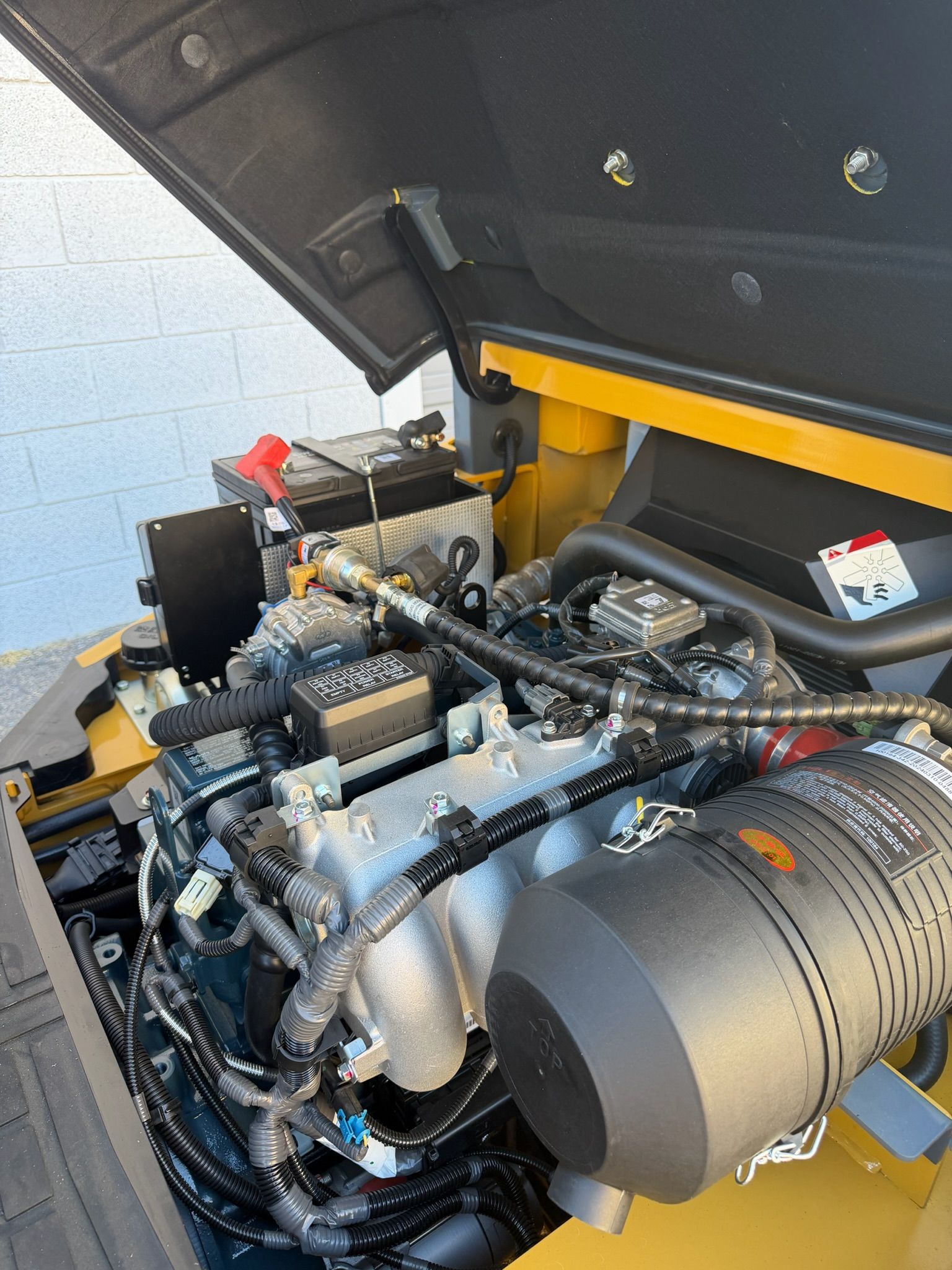 Open engine compartment with hoses, wiring, and a large gray cylindrical component under a raised hood