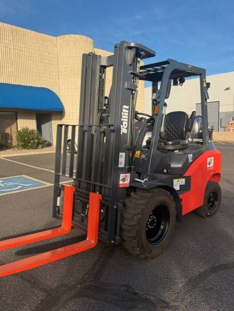 Red forklift with orange forks parked outside a beige building near a blue awning.