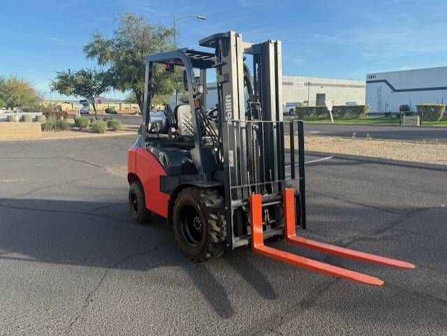Red forklift with orange forks parked on an empty asphalt lot near buildings